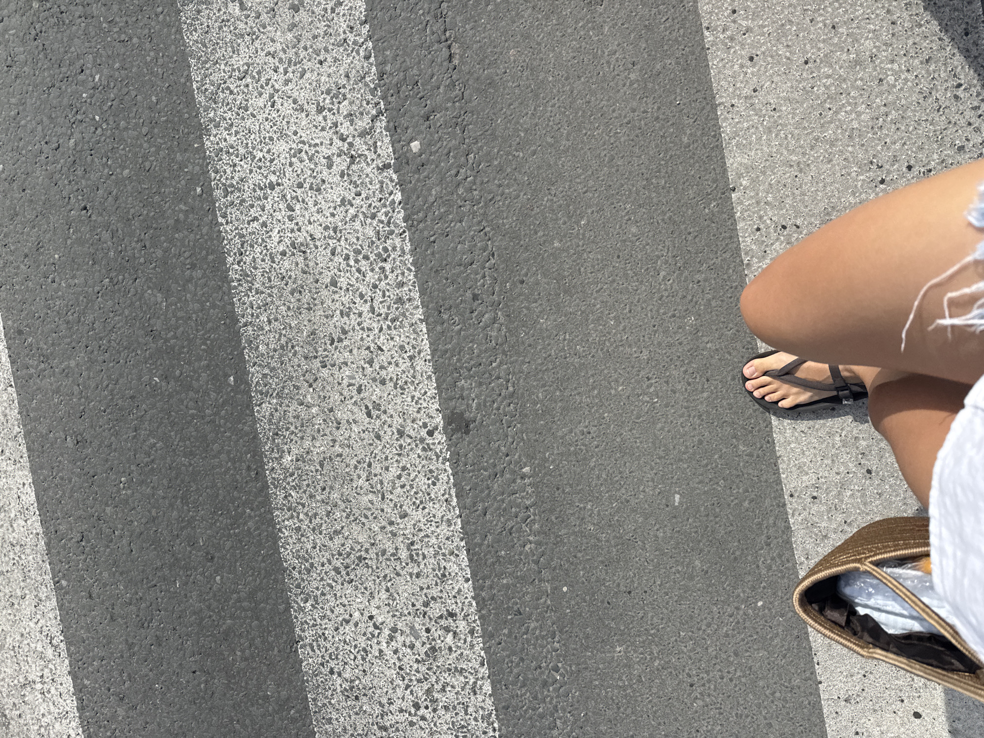 A close-up, overhead shot shows a person's legs and feet as they stand on a crosswalk. The person is wearing black flip-flops and denim shorts. A woven tote bag is visible to the left of the person's legs. The crosswalk stripes are clearly defined against the asphalt road. The lighting suggests it is a sunny day.