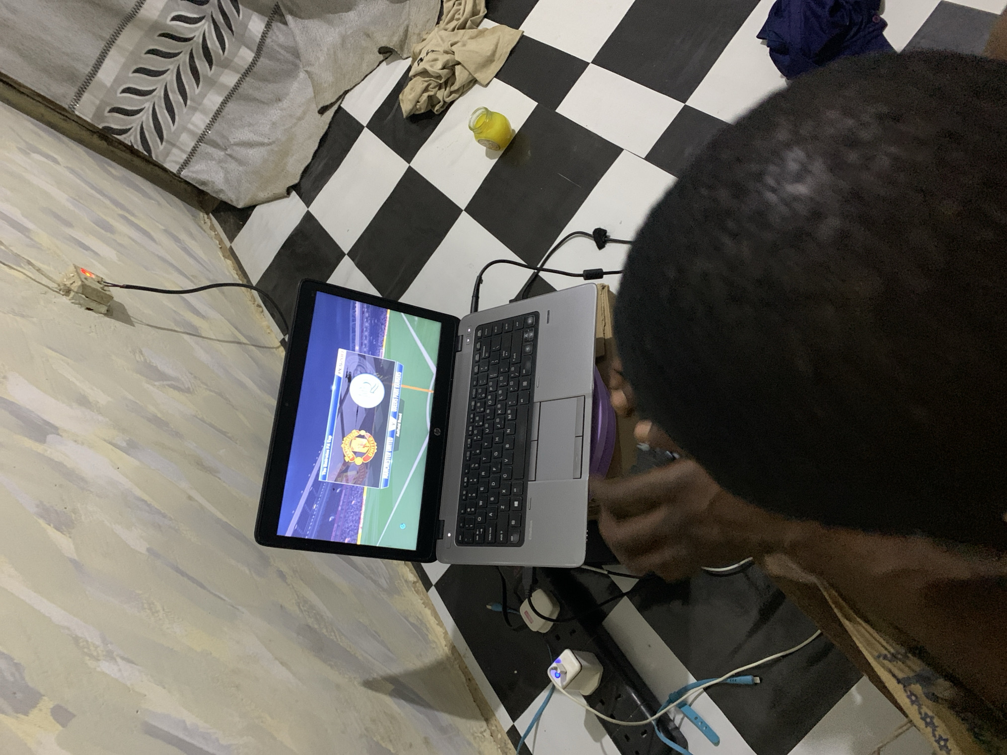 The image depicts a person in a room with a black and white checkered floor and light beige walls. The person is looking at a laptop computer, which is displaying a soccer game. The laptop is connected to a power source via a black power cord, which is plugged into a white outlet on the wall. There is a power strip plugged into the wall outlet with multiple other plugs. The environment contains several additional items, including a jar of liquid and cloth bundles on the floor. The image likely depicts the setting of an indoor room with a person operating a laptop, potentially for entertainment or other purposes. Rafin Par, Nigeria is not discernible from the image.