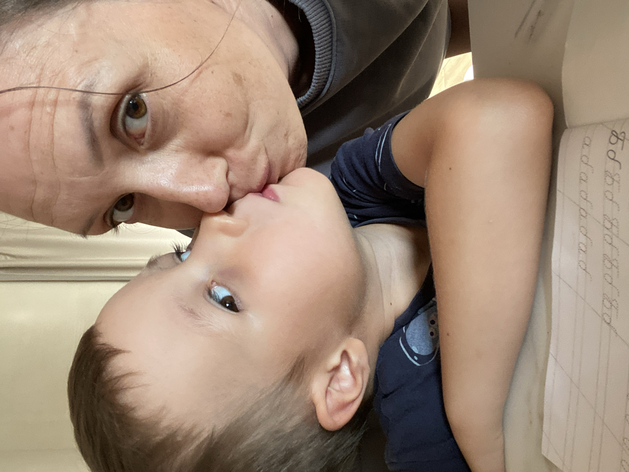 An extreme close-up, low-angle selfie shows a woman and a young boy kissing cheeks. The boy's head is tilted up towards the woman's face, with his lips touching her cheek. He has light brown hair, blue eyes, and fair skin. He is wearing a dark blue t-shirt. The woman has brown eyes, light brown hair, and fair skin with some freckles and wrinkles. She is wearing a gray sweatshirt. They are positioned over a notebook with lined paper that has cursive writing on it, suggesting they are at a table. The background is blurred, showing a neutral-colored wall and curtain, indicating an indoor setting. This scene appears to be captured in a domestic environment. The image does not contain information about the city of Chaikovsky, Russia.