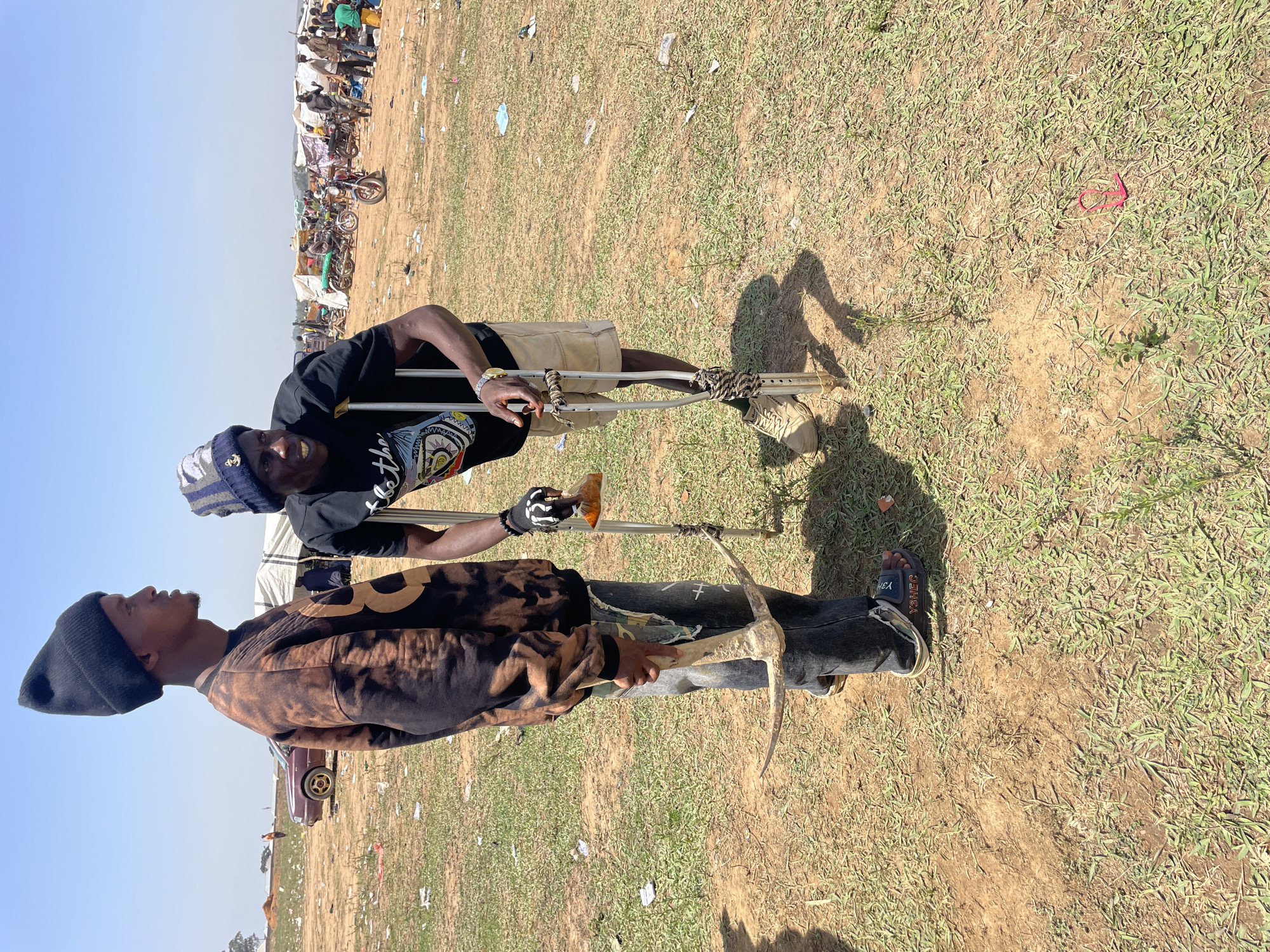 Two men stand in a grassy field strewn with trash, likely in Maiyanga, Nigeria. One man uses crutches, wearing a black t-shirt, khaki shorts, and a knit hat. The other holds a pickaxe, wearing a black beanie, a tie-dye long-sleeved shirt, and jeans. In the background, a crowd of people, tents, and vehicles is visible. The sky is blue.