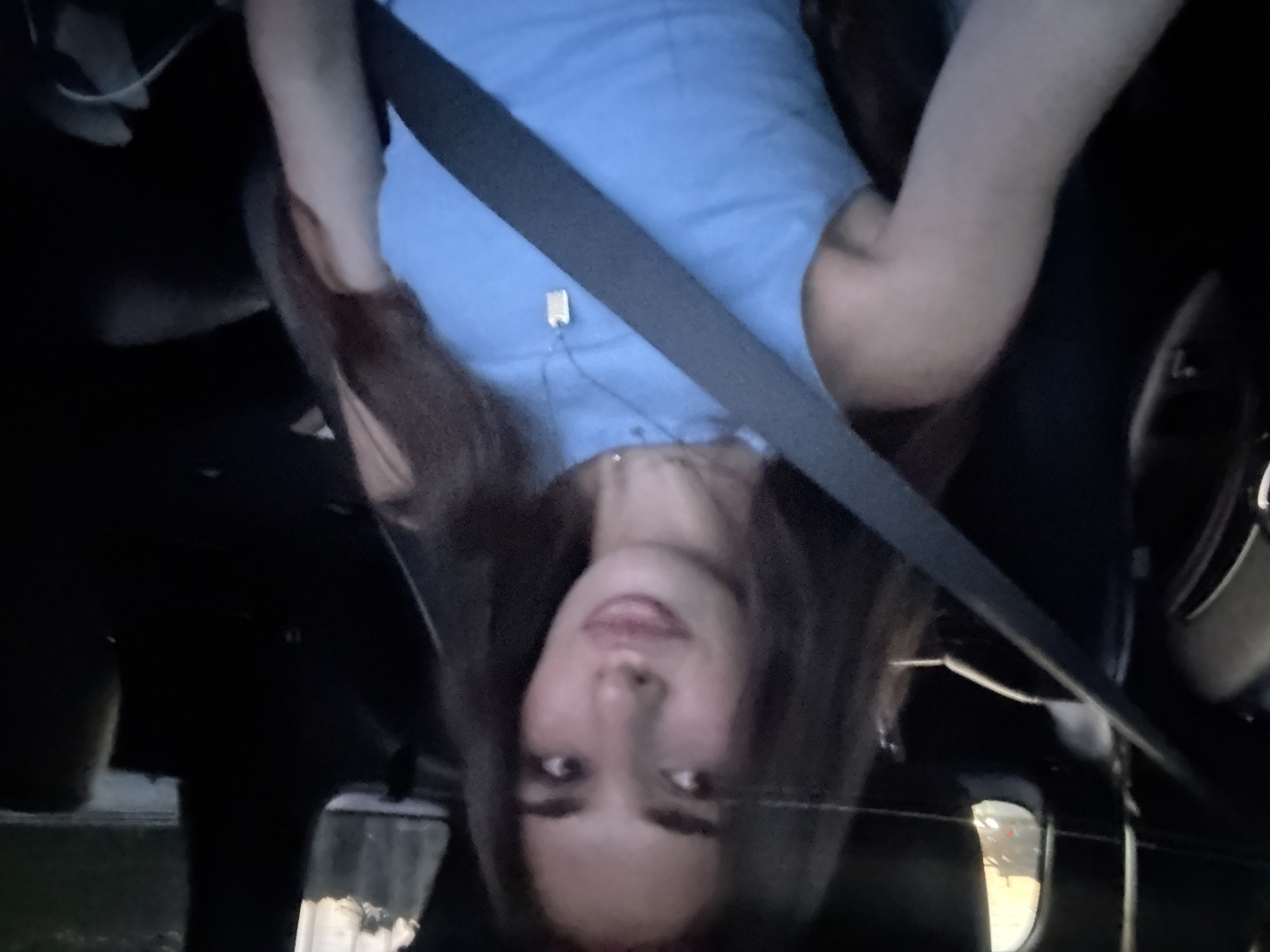 The image displays a single female individual seated in the front passenger seat of a vehicle, in what appears to be a low-light setting consistent with evening or night, within the city of Boca Raton, United States of America. The woman, with long, dark brown hair and a fair complexion, is looking directly towards the camera. She is wearing a light blue sleeveless top and a silver-colored chain necklace with a rectangular, dog tag-style pendant. A dark-colored seatbelt is securely positioned diagonally across her chest and lap. The vehicle's interior, including the seats and visible panels, is dark. Through the car windows, blurred bright lights and faint, dark environmental outlines are visible, suggesting an urban or suburban road environment with ambient light sources such as streetlights or other vehicle lights, and possibly distant structures or foliage. A yellowish-orange glow is faintly visible through the upper portion of the driver's side window.