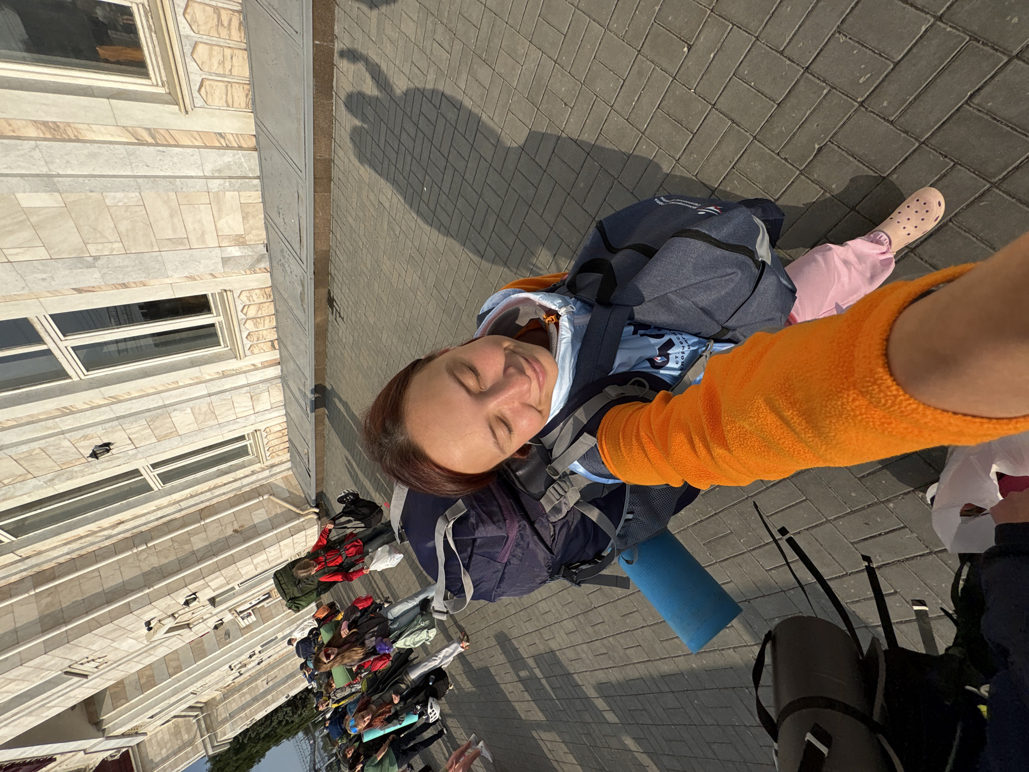 A low-angle, outdoor, eye-level shot features a young woman with her eyes closed, smiling, as she holds a backpack and is being filmed, possibly as part of a travel vlog. The woman is wearing a light blue hooded jacket over a dark blue shirt, and pink sweatpants. She has an orange sleeve visible, indicating a layered outfit. She is carrying a large blue and grey backpack, with a blue yoga mat strapped to it. In front of her, a black duffel bag is visible on the ground. The background shows a large building with beige and white marble-like facade, featuring tall windows and arched decorative elements. A group of people, some with backpacks, are walking in the background towards the building. The ground is paved with grey rectangular stones. The scene appears to be in front of a train station or a similar public building. The lighting suggests it is daytime, with long shadows cast on the ground. Based on information associated with the building's architectural style, this scene is likely located at the Izhevsk Railway Station in Izhevsk, Russia.
