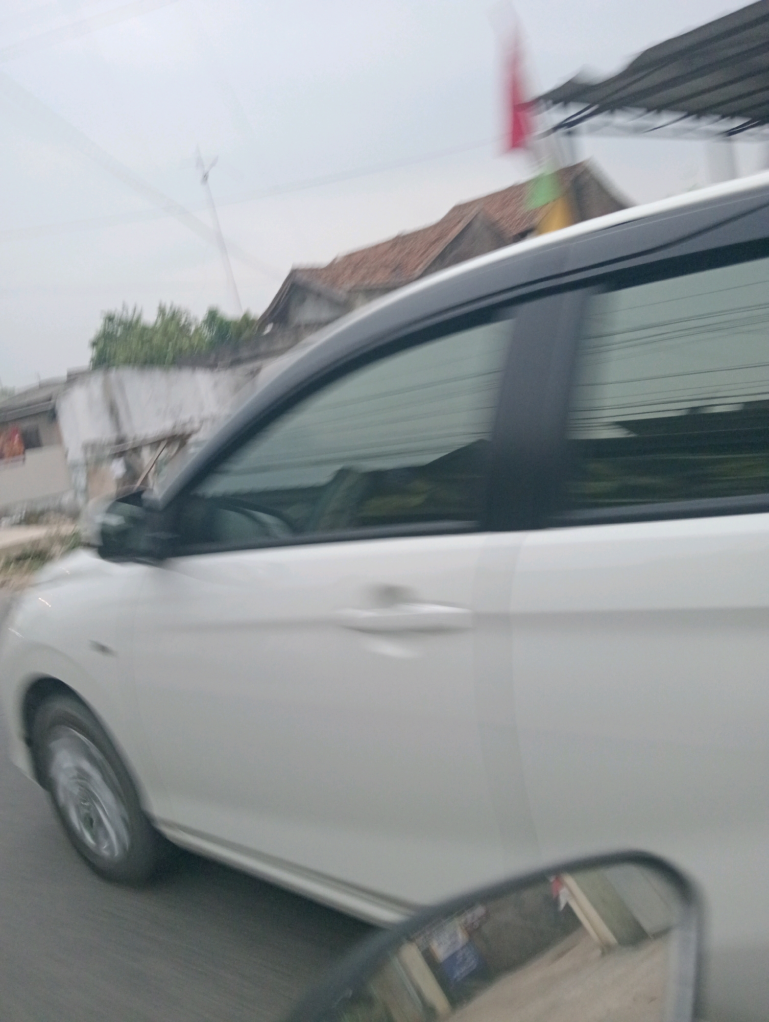 The image shows the side of a white car in motion, with the focus on the driver's side door and window. The car is captured with motion blur, suggesting it is traveling. In the background, there are buildings with reddish-brown tiled roofs and some greenery. A blurred red flag is visible on the right side of the image. The sky is overcast. The image appears to have been taken from another vehicle. The scene is consistent with a street view in Pringsewu, Indonesia, given the architectural style and vegetation.