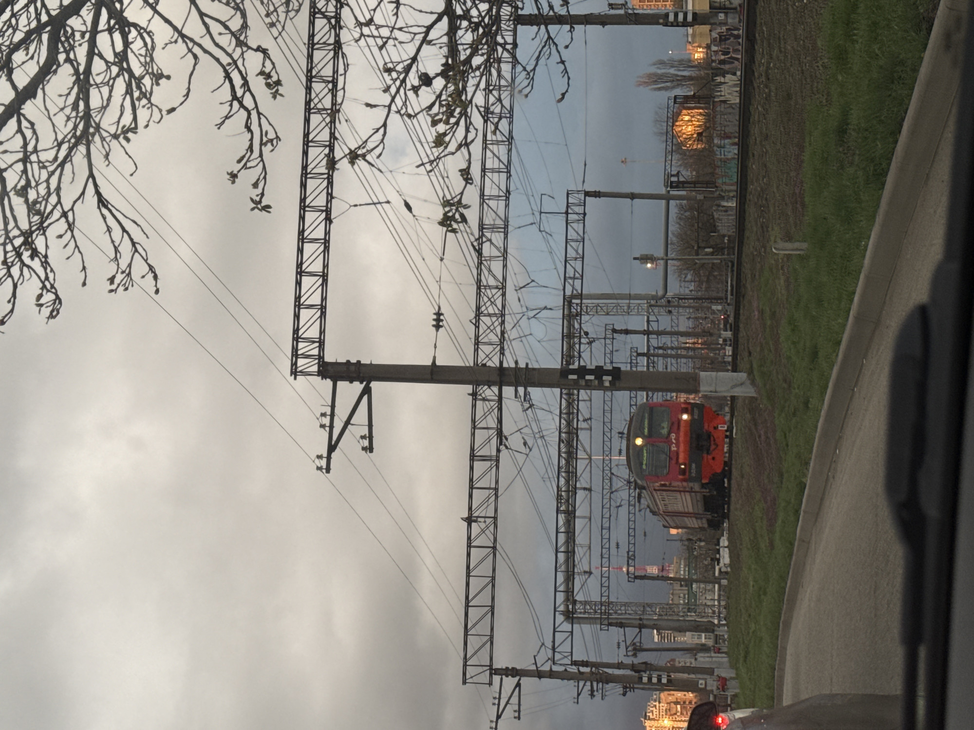 The image presents a view of a red passenger train traveling on a railway track, with a network of metal power line structures and overhead wires dominating the foreground and center. The train is positioned to the right side of the frame, with its front illuminated by headlights.
The backdrop features a cityscape, including high-rise buildings. In the upper portion of the frame, tree branches are visible against a cloudy sky, and to the right, there is greenery and a road. The composition is shot from within a vehicle.
The location is unknown, but the urban environment suggests a city setting.