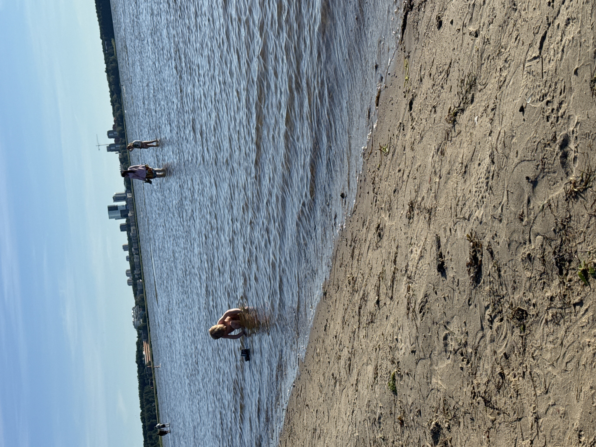 The image depicts a sunny day at a beach in Tallinn, Estonia, likely near the Pirita district, as evidenced by the cityscape and coastline visible in the background. The foreground is dominated by a sandy beach with scattered seaweed and debris. Gentle waves lap the shore of a large body of water, which appears to be the Baltic Sea.
In the water, a young child is crouched down, playing with a toy truck near the shoreline. Further out in the water, a woman wearing a pink top and a child in a light-colored swimsuit are standing. In the distance, several other people are walking along the water's edge or wading. The background features a tree-lined shore and a cluster of modern buildings, including high-rises and cranes, characteristic of urban development in Tallinn. The sky is clear and blue, with a bright sun casting a warm glow over the scene.