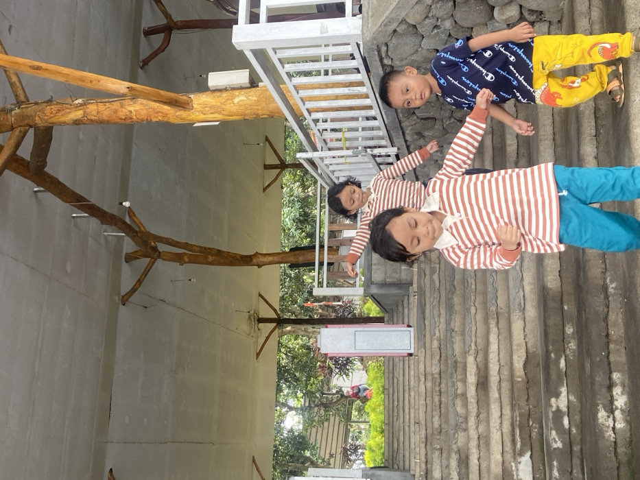 This outdoor image captures three young children in a recreational or garden setting in Durensewu, Indonesia, during the daytime.
In the foreground, a child with dark hair is dressed in a brown and white striped long-sleeved shirt and blue pants, exhibiting a joyful expression with arms slightly raised, suggesting movement or play. Directly behind this child, another child in an identical striped shirt and blue pants is also visible, appearing to be engaged in the same activity. Further back and to the right, a third child, a boy, stands on a series of concrete or stone steps. He wears a blue t-shirt with the word "Champion" prominently displayed in white text, paired with bright yellow patterned pants. This boy looks directly towards the viewer with a neutral expression.
The children are positioned on or adjacent to ascending stone steps that occupy the right side of the frame. To the left, a structure incorporates large, natural tree branches or logs, which serve as decorative or structural elements, possibly supporting a covered area or balcony. A white metal railing is visible above these wooden features. The background reveals lush green foliage and trees, indicating a natural environment. A grey and pink box-like object stands near the steps in the mid-ground. The lighting is bright and diffused, characteristic of daytime. The overall scene conveys a playful and relaxed atmosphere.