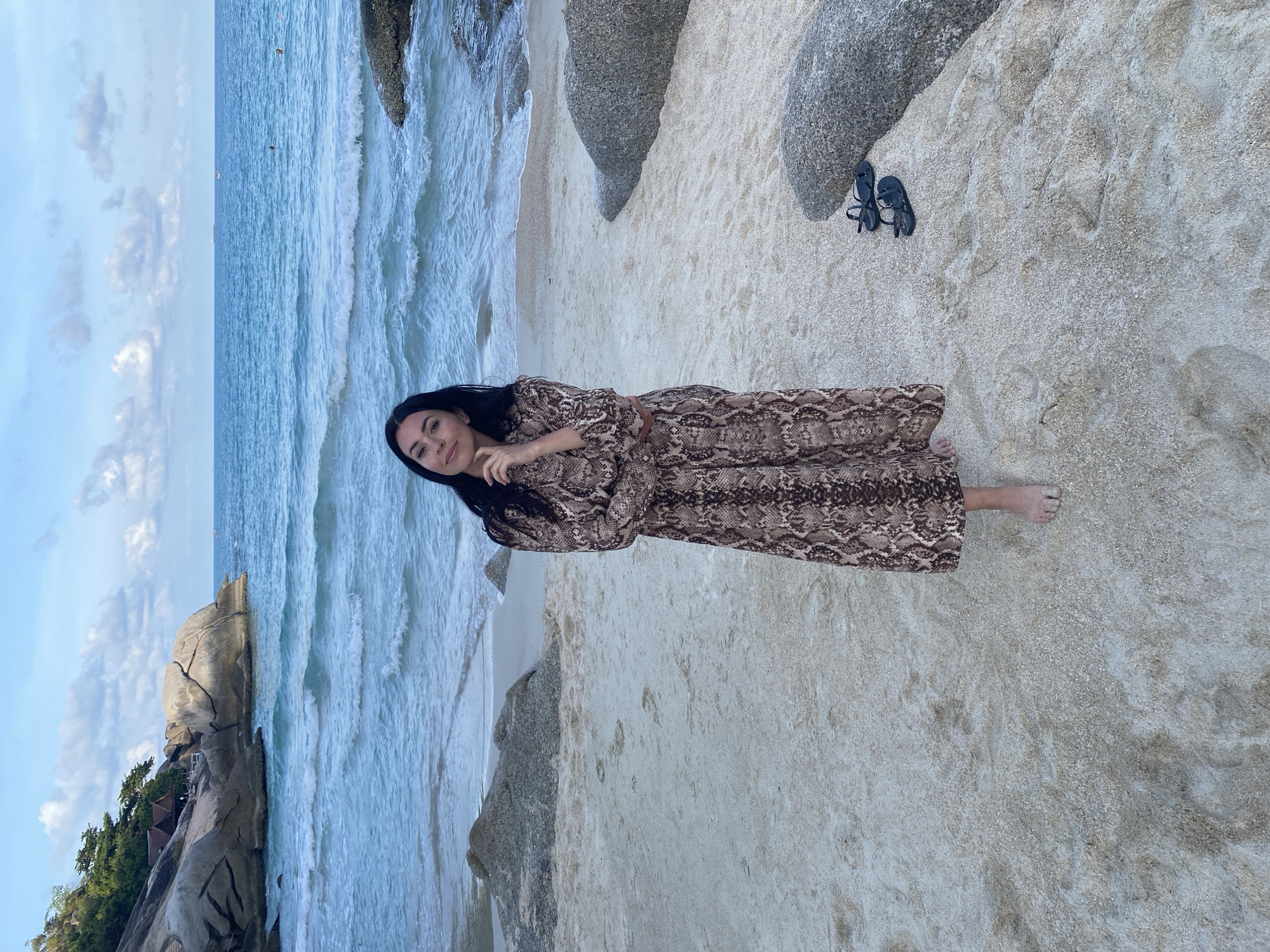A woman in a long brown patterned dress stands on the sandy shore of Ban Haad Rin, Thailand, near large rocks and the ocean. Her black sandals are placed on the sand near the rocks.