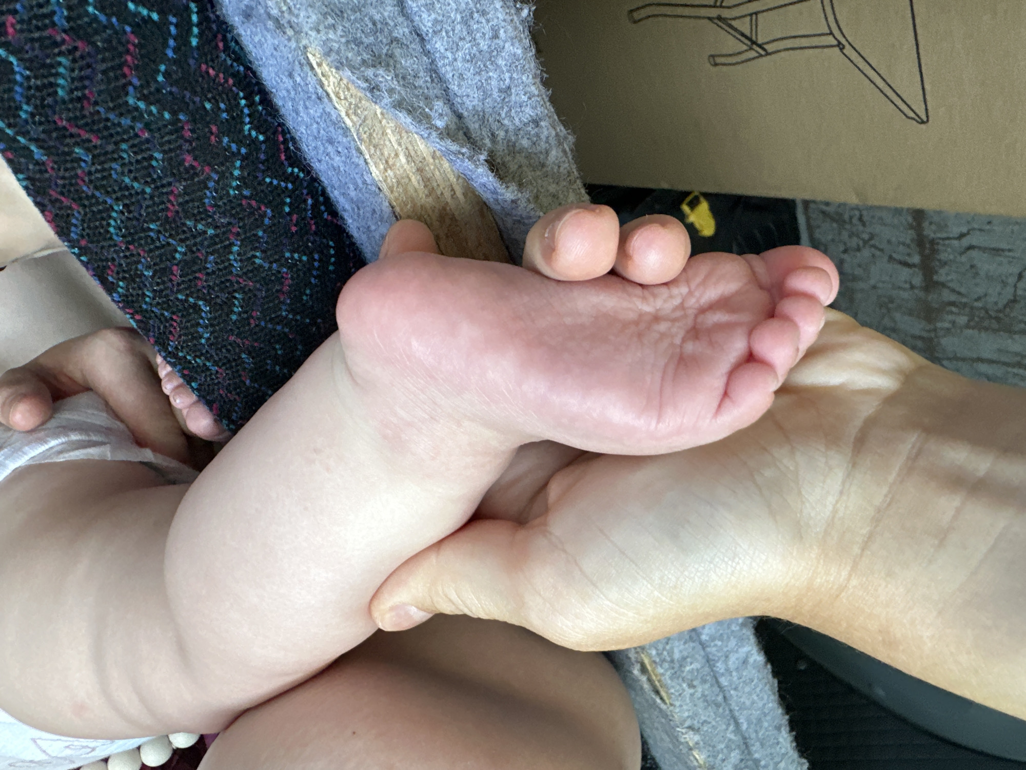 An adult human hand is holding the foot and leg of a baby or toddler. The baby is wearing a diaper. The background contains fabric with multicolored designs and a cardboard box with a chair drawing. The location is not discernible from the image.