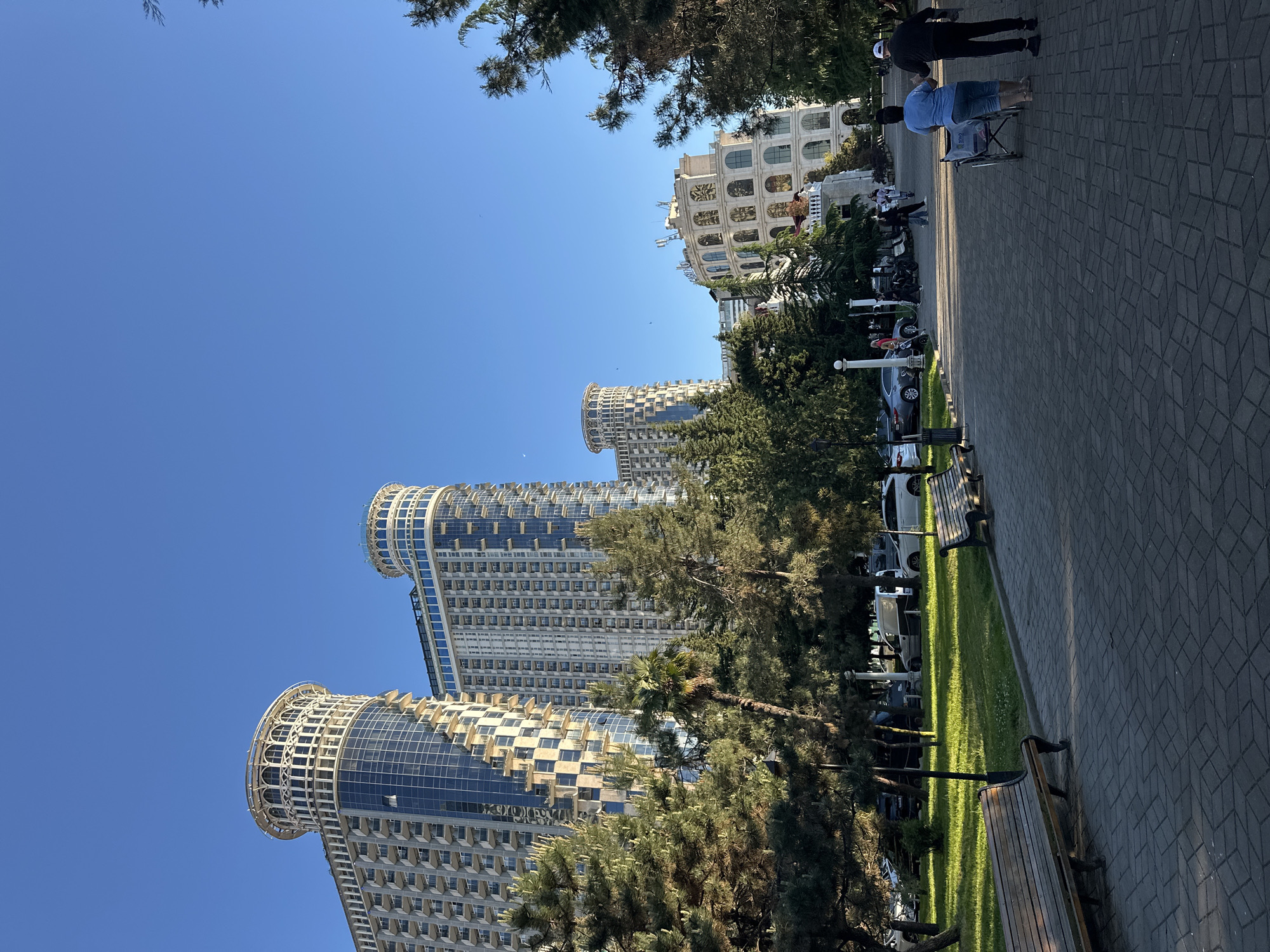 Captured from a low-angle perspective, the image showcases the skyline of Batumi, Georgia, under a clear blue sky. Dominating the scene are three modern, high-rise cylindrical buildings with distinctive architectural designs, possibly hotels or residential complexes, partially obscured by lush trees. The buildings appear to have a complex structure with numerous windows and curved balconies.
Adjacent to the buildings is a paved walkway or park area with benches, a neatly maintained strip of grass, parked cars, and street lamps. In the background, a building with a classical architectural style can be seen. On the walkway, a few individuals are visible, including a person in a wheelchair accompanied by another person pushing it. The scene evokes a sense of urban development and a pleasant environment for leisure activities.