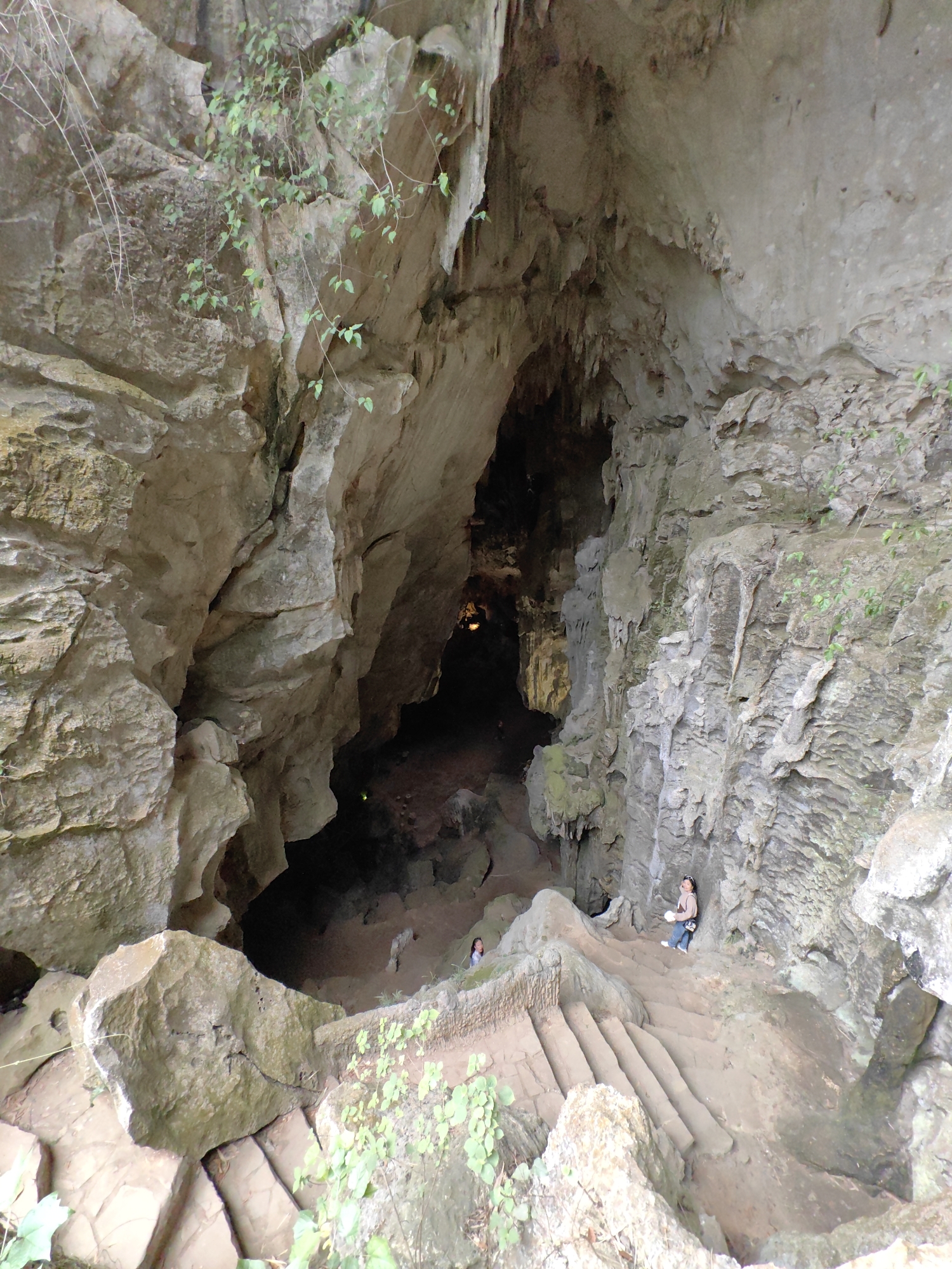 Here is a detailed description of the image:
The image shows the entrance of the Tam Thanh Cave in Lạng Sơn, Vietnam. The view is from above, looking down into the cave's opening. The cave entrance is surrounded by large, jagged limestone rocks. There are steps leading down into the cave. Two people are visible on the steps, and another person is inside the cave closer to its interior. Some plants are growing on the rocks near the steps. The interior of the cave is dark and dimly lit with some lighting within, with visible rock formations, presumably stalactites and stalagmites.