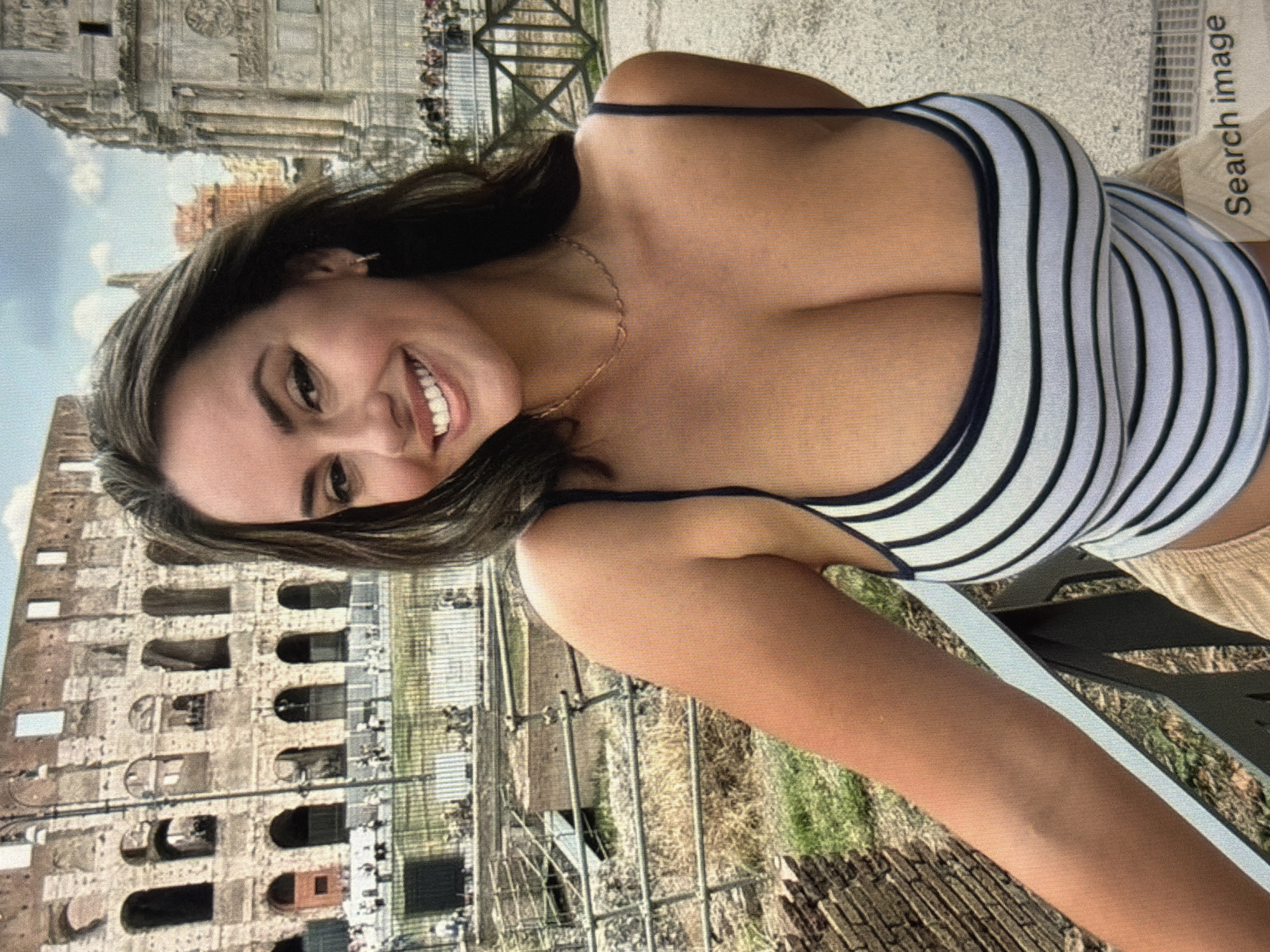 A woman is pictured in front of the Colosseum in Rome, Italy. She is wearing a striped tank top and tan shorts. The Colosseum is a large, ancient amphitheater with multiple levels of arches. The woman is smiling and looking at the camera. The background shows a bright, sunny day with some clouds in the sky.