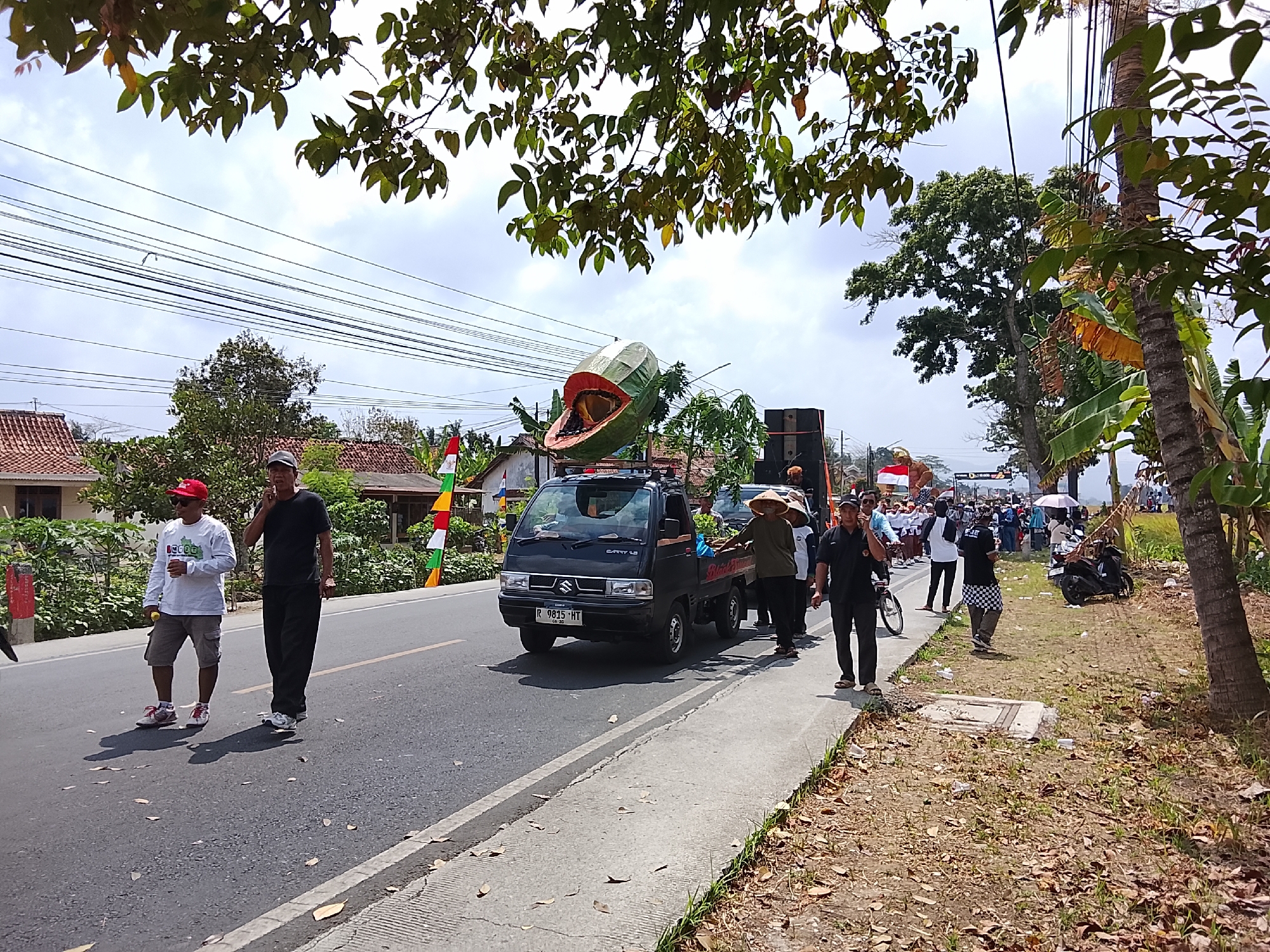The image depicts a street scene in Sampang, Indonesia, likely during a parade or festival. The foreground shows a paved road with a concrete sidewalk. Two men walk on the road, one wearing a red cap and white long-sleeved shirt, and the other in a black t-shirt and black pants, talking on a phone. A black Suzuki Carry pickup truck is driving on the road, carrying a large, inflatable object resembling a sliced fruit, possibly a papaya, mounted on its roof. Several people are walking alongside the truck and on the sidewalk. One person wears a conical hat. Flags, including the Indonesian flag, are visible on poles. The background features trees, houses with red tiled roofs, and utility poles with electrical wires. The sky is overcast. The overall atmosphere suggests a public event.