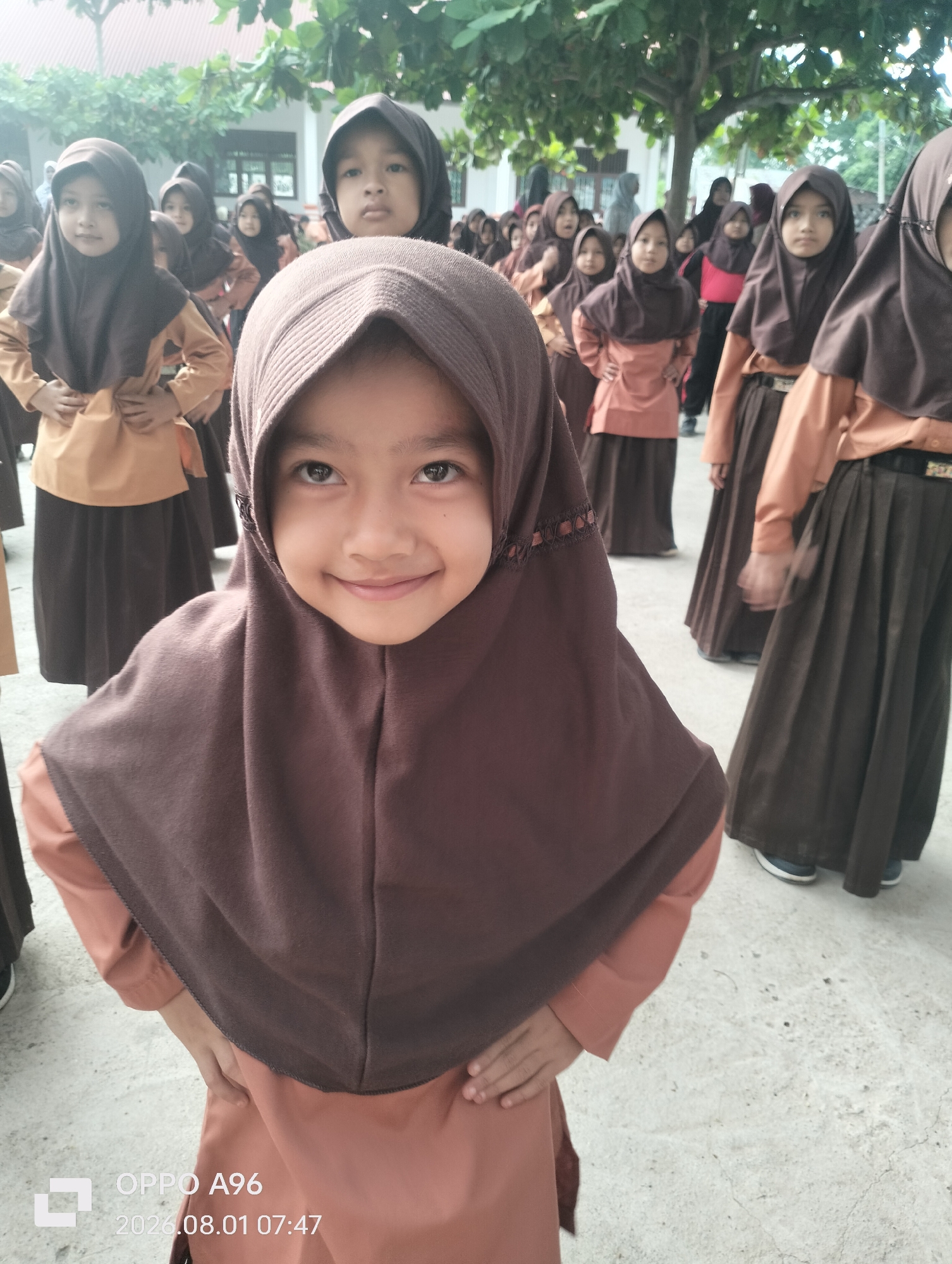 In Keudekarieng, Indonesia, a group of young girls, dressed in similar brown hijabs and peach-colored uniforms, stand in an outdoor schoolyard. The girl in the foreground, with her head tilted slightly and a wide smile, is the central focus. Her dark eyes are directed towards the camera, and her hands are resting on her hips. Behind her, other girls are visible in varying degrees of focus, some standing with their hands clasped in front of them and others looking in different directions. The background suggests a school environment, with trees and a building visible. The ground appears to be a paved surface, possibly concrete. The image is a candid shot, capturing a moment of everyday life at the school.