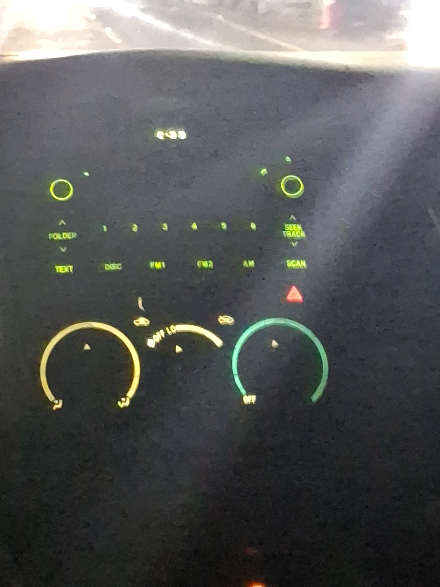 The image is a close-up shot of a car's dashboard display, likely an older model, showing various buttons and indicators illuminated in green and yellow. The display features labels such as "FOLDER," "TEXT," "DISC," "FM1," "FM2," "AM," "SCAN," and "SEEK TRACK." There are also numerical indicators from 1 to 5. Two circular illuminated elements are visible, one in yellow and one in green, resembling volume or tuning knobs. A small red triangular warning light is also illuminated. The background is dark and appears to be the interior of a vehicle. The lighting suggests it is either night or the car is in a dimly lit area.
There is no information within the image or its crops that could place it in Lumajang, Indonesia, or any specific city. The image is purely of a car's dashboard.