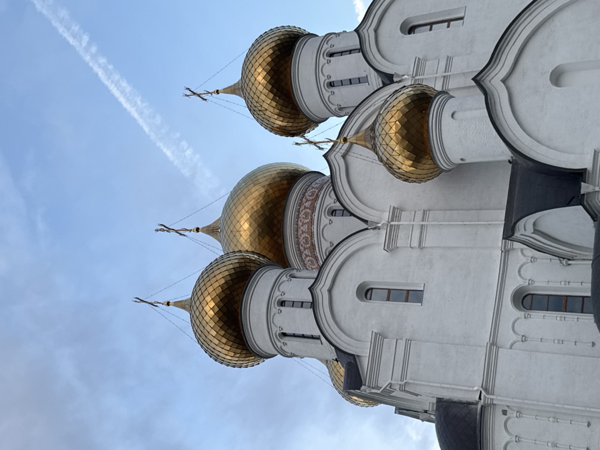 A low-angle, full shot captures the white facade and golden domes of a large, ornate church against a pale blue sky with thin white clouds and a contrail. The architecture features multiple domes, each topped with a cross. The church's white walls are decorated with architectural details like arched windows and decorative cornices. The gold domes are multifaceted and reflect the sky. The scene is devoid of people or any other activity.
