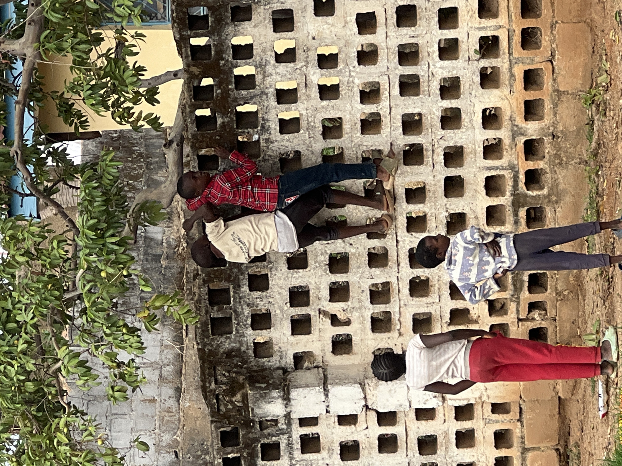 Four children are positioned along a concrete block wall in Jos, Nigeria. The wall, which is off-white and worn, has numerous square openings in its construction. From top to bottom: a child in a red plaid shirt and jeans; a child in a light-colored shirt and dark shorts; a child in a white and blue striped shirt and jeans; and a child in a white t-shirt and red pants. To the left is a tree with green foliage. The background shows a yellow building behind the wall, with a blue window frame. The environment appears to be outdoors, adjacent to the wall.