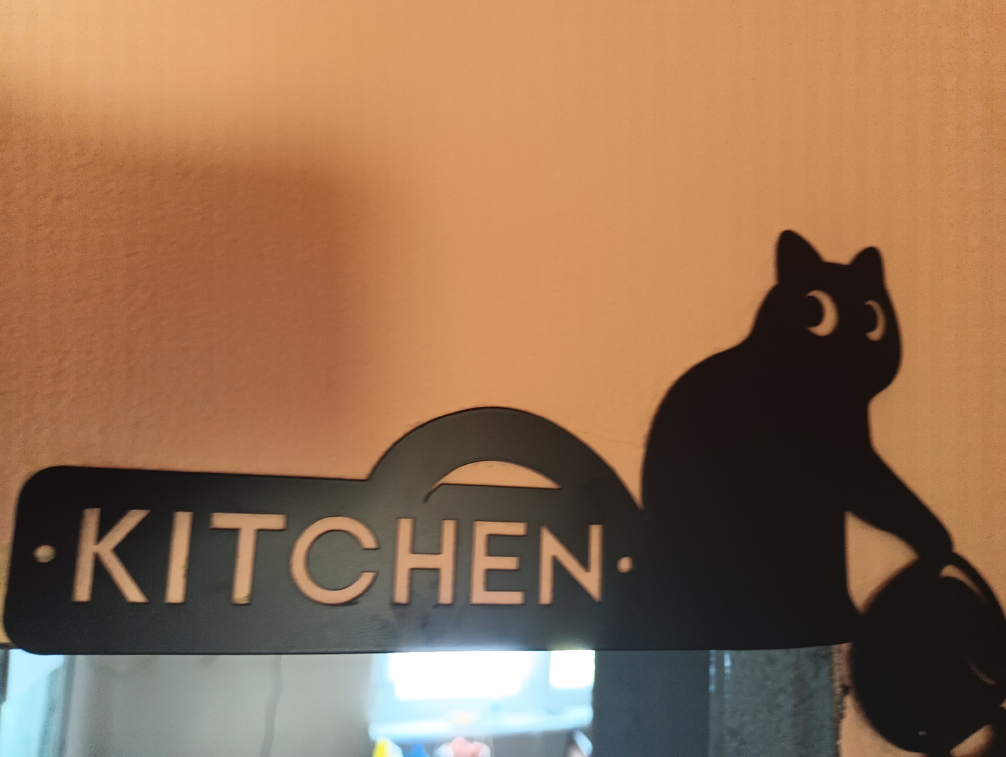 A black metal cutout of a cat is affixed to a wall. The cutout reads "KITCHEN" in cutout letters and depicts a cat to the right of the text. The cat appears to be holding a round object. The wall is a light orange or peach color. The scene is indoors, and a blurred reflection of what appears to be a room with lights is visible at the bottom of the image, suggesting it may be a sign for a kitchen. The provided location information states the image is from Karnobat, Bulgaria.