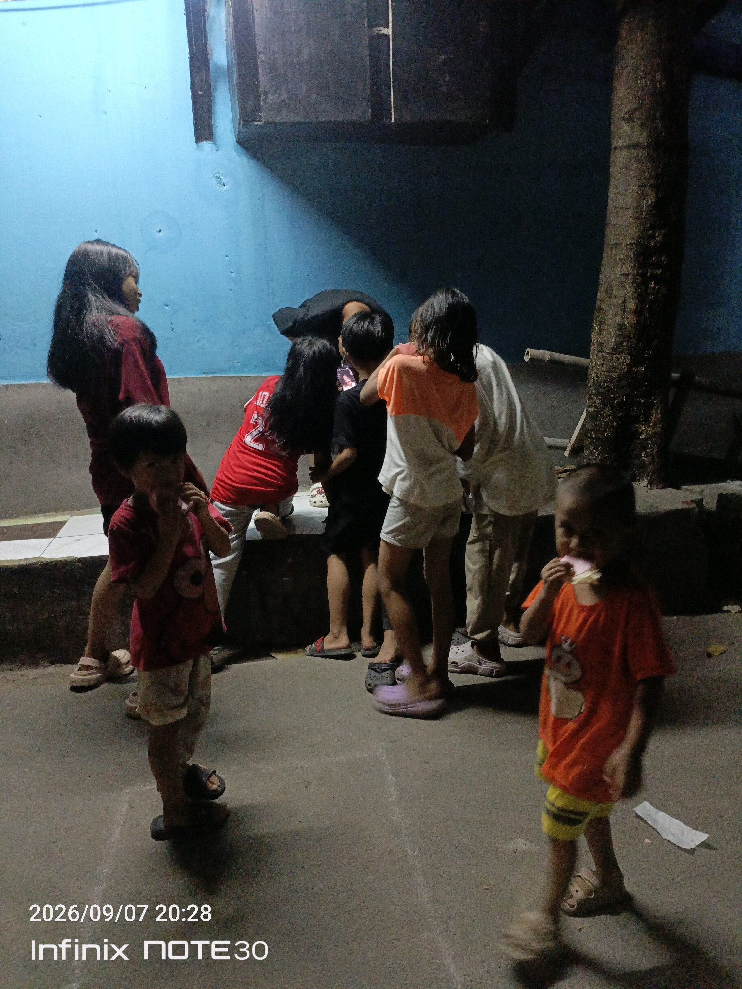 In East Jakarta, Indonesia, at approximately 8:28 PM on September 7, 2026, a group of children are gathered outdoors. The scene is dimly lit, with a blue wall forming the background on the left and a textured tree trunk on the right. Several children, appearing to be of varying ages, are positioned around the frame. In the foreground, a young child in a red shirt and light-colored shorts is holding something to their mouth, seemingly eating. Another child in an orange t-shirt and yellow shorts is also in the foreground, walking and eating a light-colored food item. A group of older children and an adult are in the middle ground. One child is wearing a red shirt with the number "22" on the back, and another is in a white and orange t-shirt. The ground is a textured, grayish surface, possibly concrete or asphalt, with some white chalk markings visible, suggesting a play area. The overall impression is a casual outdoor gathering of children in a residential or community setting.