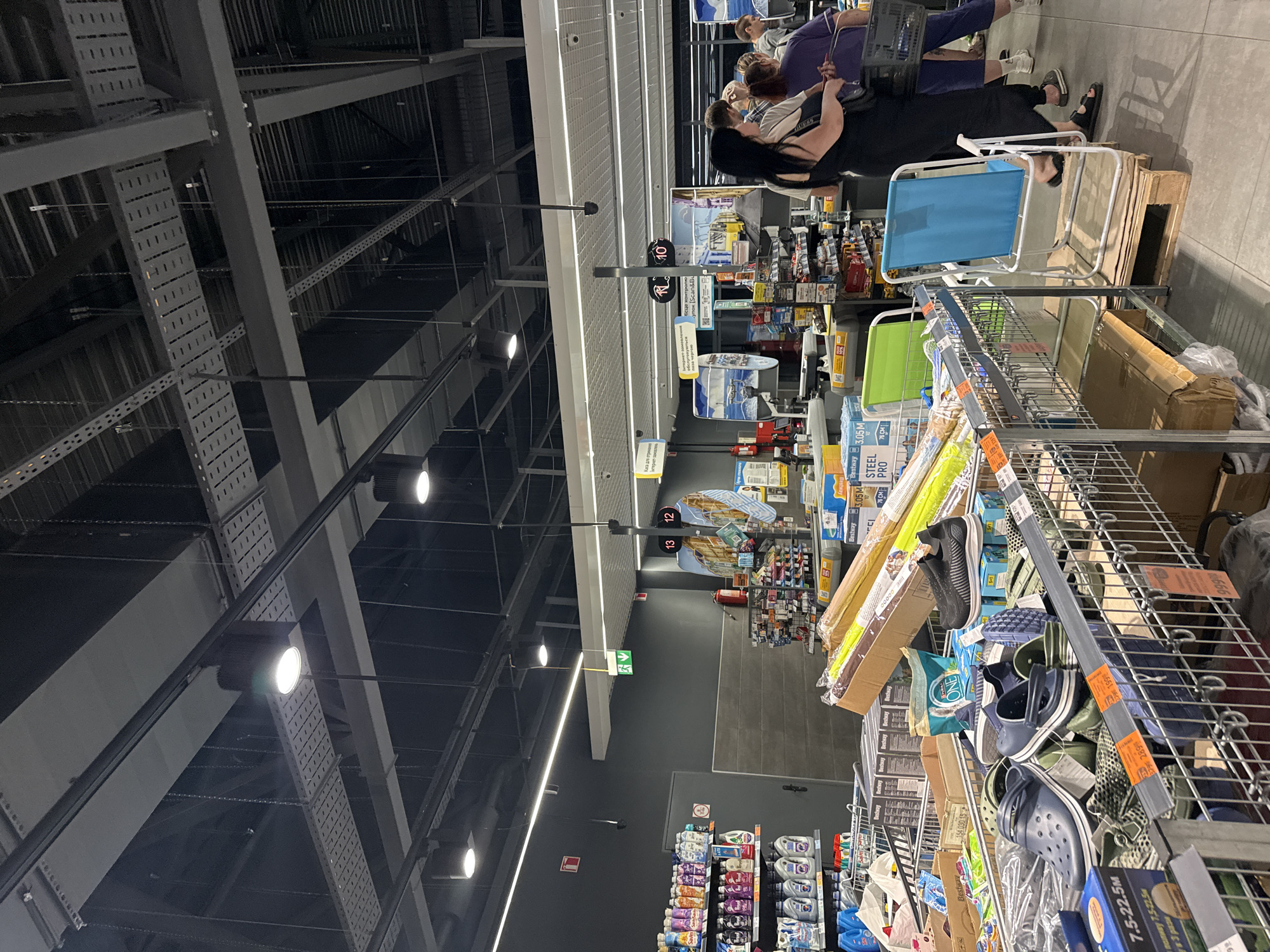 The image depicts an interior scene of a supermarket in Dnipro, Ukraine, likely a section featuring seasonal or outdoor items, as suggested by the presence of foldable chairs, pool accessories, and shoes resembling sandals or clogs. The environment is brightly lit with overhead fluorescent and LED lighting.
In the foreground, several shopping carts are filled with merchandise. One cart prominently displays blue and green clogs, along with patterned fabric items and boxed goods. Another cart holds long, rolled items that appear to be pool noodles or similar recreational equipment, alongside cardboard boxes. A blue and white foldable chair is placed on a wooden pallet to the right of the carts.
In the mid-ground, a checkout area is visible with multiple customers and staff members. A woman in a black dress is standing in line, holding a black handbag. Another person in a purple top is visible in the background, and other shoppers are standing at the checkouts. Several signs and displays are present, including price tags and promotional materials. Numbers "13", "12", and "10" are visible on illuminated displays, possibly indicating checkout lane numbers or product codes.
In the background, shelves are stocked with various cleaning supplies and other household goods. A fire extinguisher is mounted on the wall. The overall impression is a busy retail environment during a shopping day.