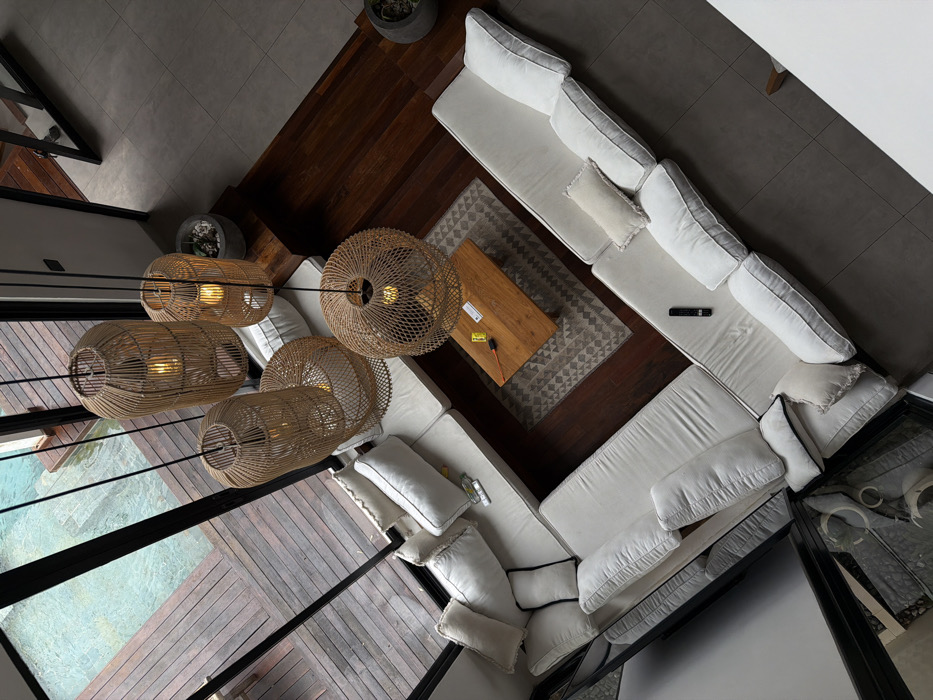 An overhead shot reveals a modern living space. A large, white sectional sofa anchors the room, positioned on a dark wood floor adjacent to a patterned rug and a wooden coffee table. Above, four distinctive woven light fixtures hang at varying heights. Floor-to-ceiling windows offer a view of a wooden deck and a pool of water, partially visible below. Neutral tones dominate the palette.