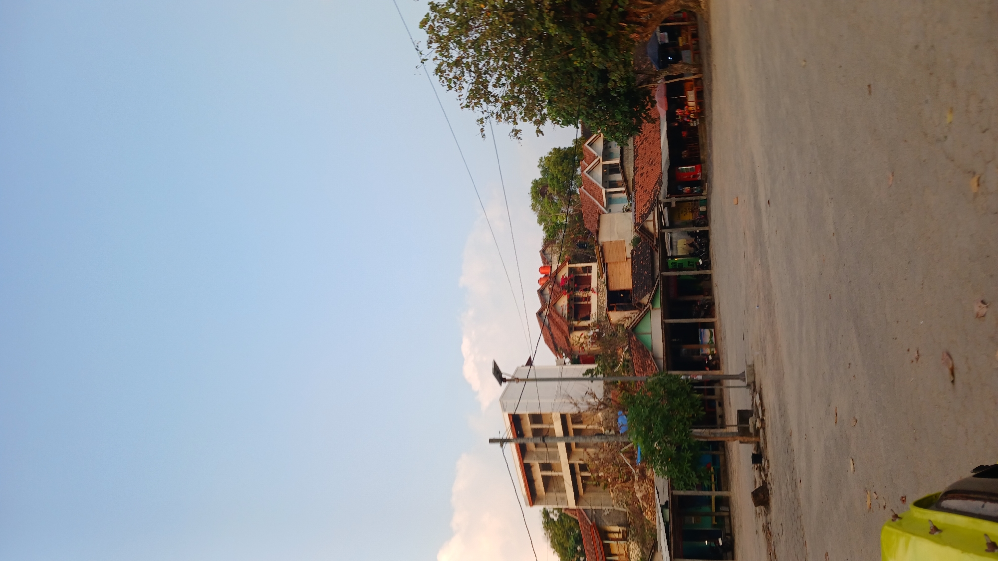 The image shows a street scene in Tepus, Indonesia, with a row of small buildings on the right and a multi-story building on the left. The sky is clear and blue with a few clouds. The buildings appear to be shops or residences, with tiled roofs and open-fronted structures. A tree is visible on the right side of the image, and power lines run across the sky. The foreground is a dusty, open area, possibly a road or parking lot, with fallen leaves scattered on the ground. A bright yellow object, possibly part of a vehicle, is visible in the lower left corner. The scene appears to be set in a rural or semi-urban area, given the simple architecture and the natural elements present.