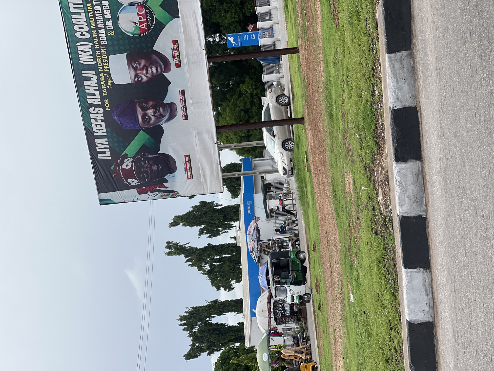 In Jalingo, Nigeria, a large billboard stands prominently above a street. The billboard displays three men in formal attire, with text in English and possibly other languages. The men are identified as "HIS EXCELLENCY BOLA AHMED TINUBU GCFR", "NENTAWE GOSHWE YILWADA NATIONAL APC CHAIRMAN", and "HIS EXCELLENCY DR. AGBU KEFA GOVERNOR TARABA STATE". Below these individuals is the text "ILIYA KEFAS ALHAJI (IKA) COALITION" and "FOR TARABA NORTH HALIN MUTUM JIGA SUPPORT PRESIDENT BOLA AHMED TI & DR. AGBU". An "APC CHANGE" logo is also visible.
In the foreground, a street with a black and white painted curb is visible. Beyond the curb, a patch of green grass transitions to brown soil. To the left of the billboard, a white building with a blue stripe features a "Union Bank" sign and an ATM. Several parked vehicles, including a silver sedan, a green and white motorized tricycle (Keke Napep), and a yellow tricycle, are present on the street. In front of the Union Bank, a few individuals are visible near a fence. A makeshift market with stalls covered by umbrellas is set up along the sidewalk, with people browsing and interacting. The overall environment suggests a busy urban setting with commercial activity.