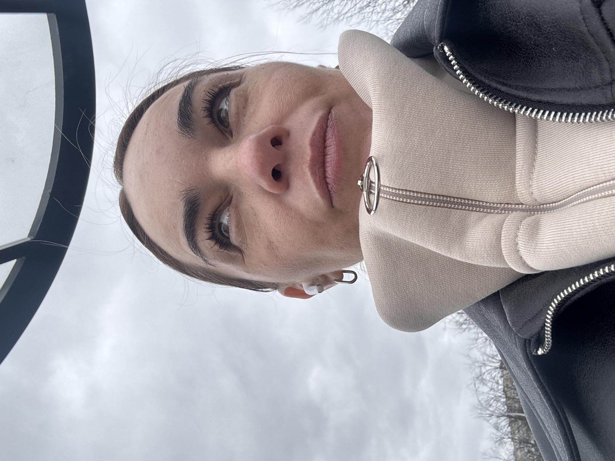 A low-angle, close-up portrait shows a woman looking directly into the camera, positioned against a cloudy sky. She wears a black leather jacket over a light beige hooded sweatshirt or hoodie, which features a silver zipper with a distinctive circular ring pull. A white wireless earbud is visible in her right ear. The woman has dark, expressive eyes and a neutral or slightly serious facial expression.
The setting is outdoors, characterized by an overcast, grey sky that fills the upper portion of the frame. Bare tree branches are visible in the upper right background, suggesting a cold season such as late autumn, winter, or early spring. On the far left, a dark, curved frame, possibly part of a structure or large object, is partially visible. The perspective implies the image is a selfie or taken from a low viewpoint, with the subject looking down towards the camera. There is no discernible activity other than posing for the photograph. No text is visible. The location context for this outdoor scene is Vsevolozhsk, Russia.
