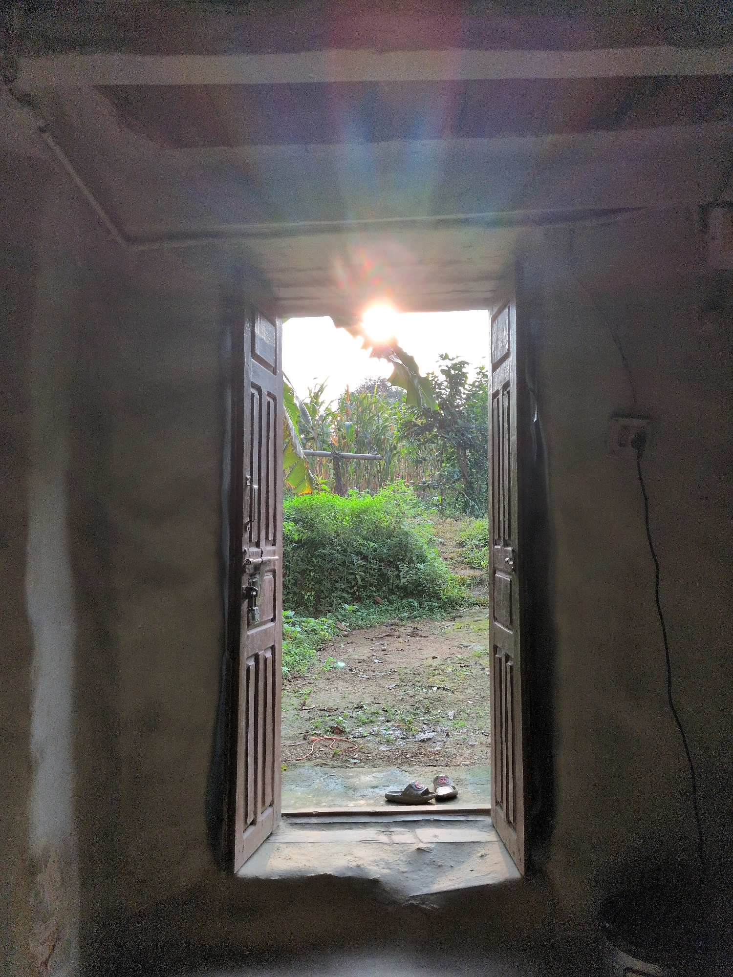 An open wooden door reveals a sunlit outdoor scene. The interior walls are rough and dark, contrasting with the bright exterior. The door frame is intricately carved, and two locks are visible on the left door. A pair of sandals rests on the threshold. Beyond the door, a dirt path leads through lush green vegetation, including banana trees and corn stalks. The sun is setting or rising, casting a warm glow and lens flare across the sky. The environment appears to be rural, likely in Tulsipur, Nepal, given the vegetation and simple structure. An electrical outlet with a cord is mounted on the right interior wall.