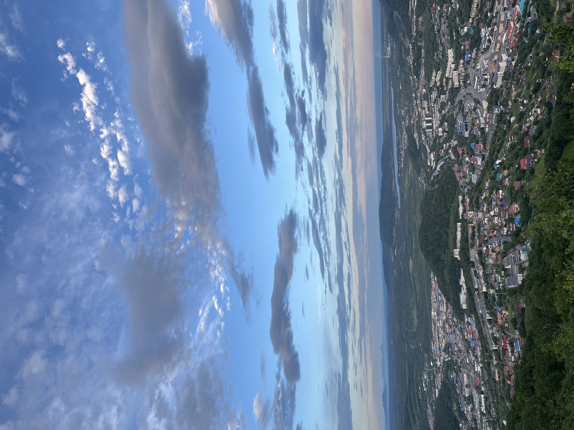 The image is a high-angle aerial view of a city nestled within a valley, likely Petropavlovsk-Kamchatsky, Russia. The city is densely packed with a variety of buildings, including residential houses with colorful roofs, apartment complexes, and some industrial structures. Roads wind through the urban landscape, with some visible vehicles. The city is surrounded by lush green, tree-covered hills, suggesting a natural and somewhat wild environment. In the distance, a body of water, possibly the Pacific Ocean or Avacha Bay, is visible, with a faint outline of mountains on the horizon. The sky is a prominent feature, filled with a mix of blue sky and various types of clouds – some wispy and white, others larger and darker gray, creating a dynamic sky. The lighting suggests it is either early morning or late evening, with soft light illuminating the scene. There are no visible people in the image.