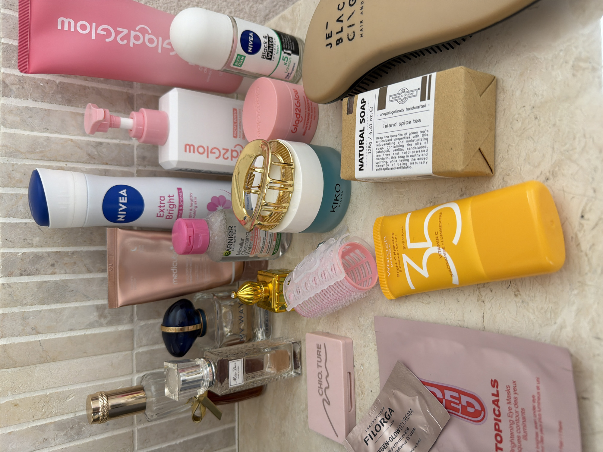 The image shows a collection of beauty and personal care products arranged on a surface made of light-colored, brick-like tiles. Items include Glad2Glow skincare products, Nivea creams and deodorant, a Kiko Milano product, a Republic of Soap bar ("island spice tea"), Wardah sunscreen, Filorga CC cream, and Topicals eye masks. Also present are various perfume bottles, including "Miss Dior" and "My Way." A hairbrush and a hair clip are included in the collection. The scene appears to be indoors, likely in a bathroom or similar space. There is no indication of a location in the city of Mengwi, Indonesia, in the image.