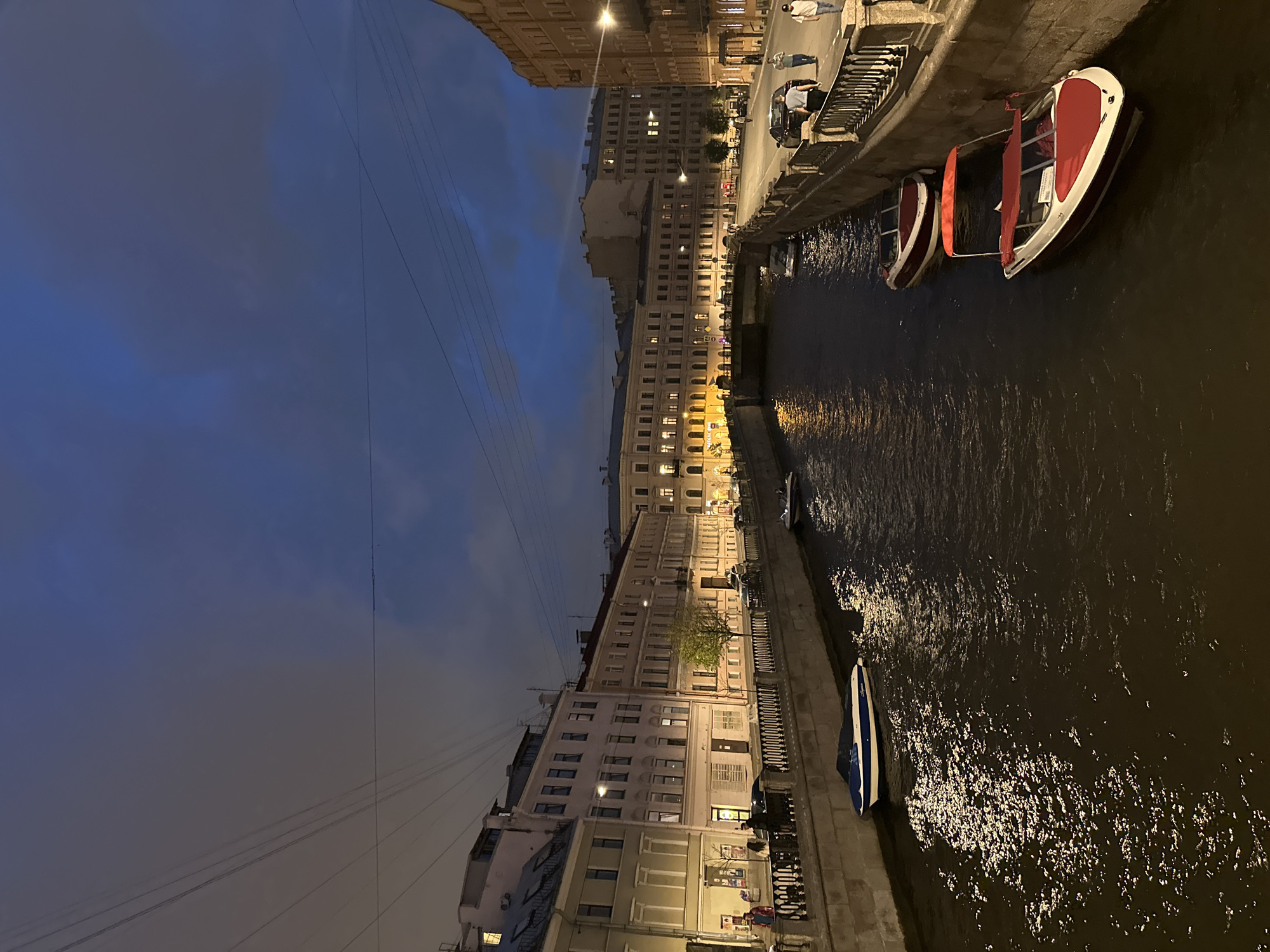 The image depicts a canal in the city of Unknown, Unknown, at night or late dusk. The sky is dark blue-grey and overcast, with several utility wires traversing the upper portion of the frame.
The left side of the canal is bordered by a stone embankment topped with a black ornate metal railing. Behind the railing, a paved sidewalk features several pedestrians and a few small trees, one with green foliage. A row of multi-story buildings, predominantly light-colored (cream, beige, light grey), lines this side, with numerous illuminated windows suggesting active interiors. A single white and blue boat with a dark cover is moored against this embankment in the foreground.
The right side of the canal also features a stone embankment with a matching black ornate metal railing. Beyond the railing, a paved sidewalk and a street are visible, with parked vehicles and several pedestrians. The buildings on this side are multi-story, generally darker-toned (brown, ochre), and also display illuminated windows. Two red and white boats are moored along this bank in the foreground; the closest boat has a red canopy. A third, smaller, lighter-colored boat is visible further down the canal near a bridge.
The canal water is dark, reflecting the artificial lights from the buildings and streetlamps, creating bright, shimmering patterns on its surface. A stone arch bridge spans the canal in the middle ground, connecting the two sides of the city.