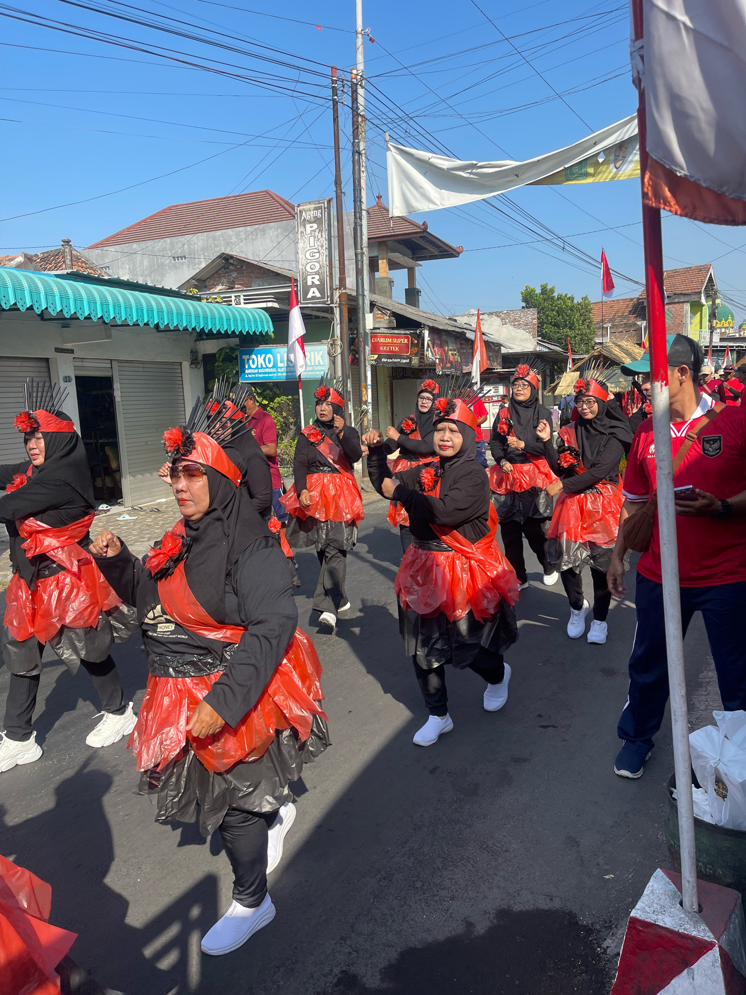 In Candi, Indonesia, a parade is in progress on a sunny day, captured from a street-level perspective. Several women, dressed in black hijabs and long-sleeved black tops, are participating in the parade. Their attire is distinctive, featuring bright red, layered, ruffled skirts made from what appears to be plastic material, worn over black leggings. They also wear matching red headpieces adorned with black and red floral accents, and some have decorative spikes extending upwards from the headpieces. One woman in the foreground, wearing sunglasses, has her right arm raised with her fist clenched. Behind her, other women are also marching, some with their arms in similar poses.
The parade is taking place on a paved street lined with buildings on both sides. Shops with awnings are visible, including one with the sign "TOKO LISRIK". Flags and banners are displayed along the street. One large white banner with some green and yellow graphics is stretched across the street overhead. In the background, a man in a red shirt is visible, facing away from the camera, and further down the street, other participants and spectators can be seen. The environment suggests a typical urban street scene in Indonesia.