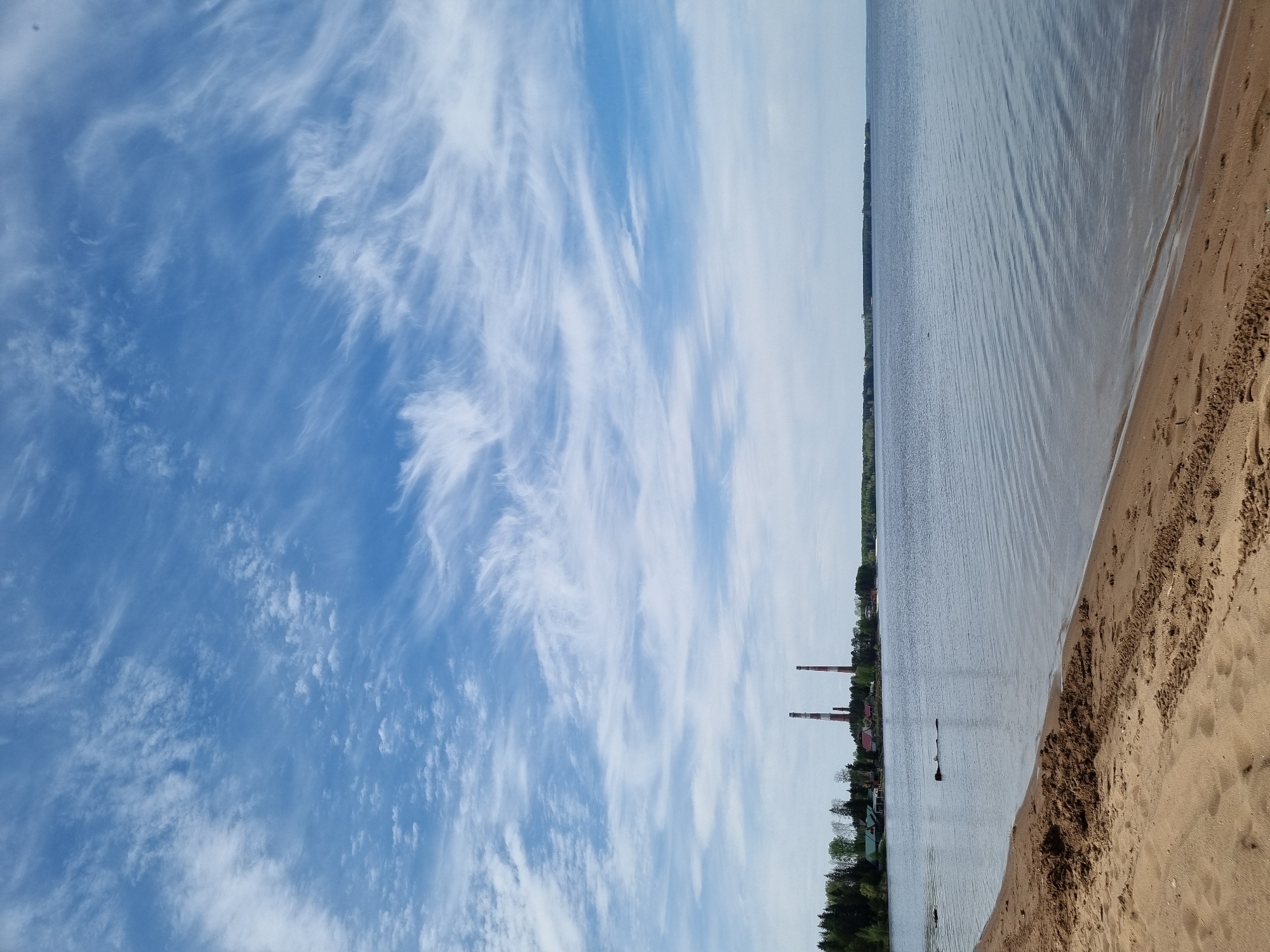 The image shows a beach in Chaikovsky, Russia, with a sandy shore and a body of water, likely the Kama River. In the background are buildings, trees, and two tall chimneys. The sky is blue with streaky cirrus clouds.