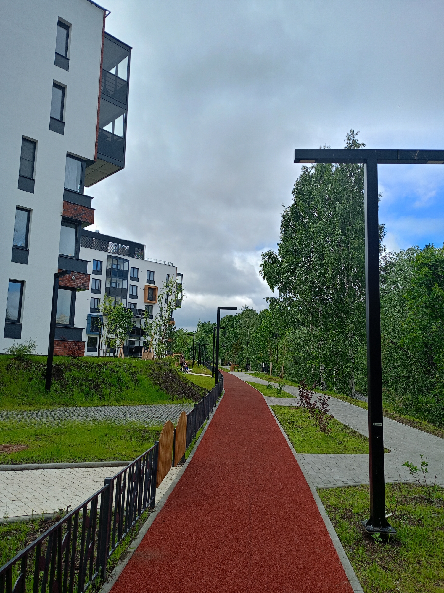 The image depicts an outdoor scene with a modern apartment building on the left and a pathway in the foreground. The building is white with dark windows and balconies, and appears to be newly constructed. Adjacent to the building is a sloped, grassy area with some young trees.
A red, cushioned pathway, likely for running or recreation, runs from the bottom right corner of the image diagonally towards the center and then curves to the left. This pathway is bordered by a light gray paved path and a black metal fence. Further along the pathway, there are modern black lampposts. Beyond the pathway, a dense line of green trees forms a natural backdrop, with a brighter sky visible through gaps in the foliage. The weather appears to be overcast, with patches of blue sky.
In the mid-ground, on the grassy area near the apartment building, two figures, a child and an adult, are visible walking. A small wooden fence segment is also present near the red pathway. The overall setting appears to be a residential complex or a park area integrated with housing. There is no visible text in the image. The time of day seems to be daytime. The scene evokes a sense of modern urban living combined with green spaces.