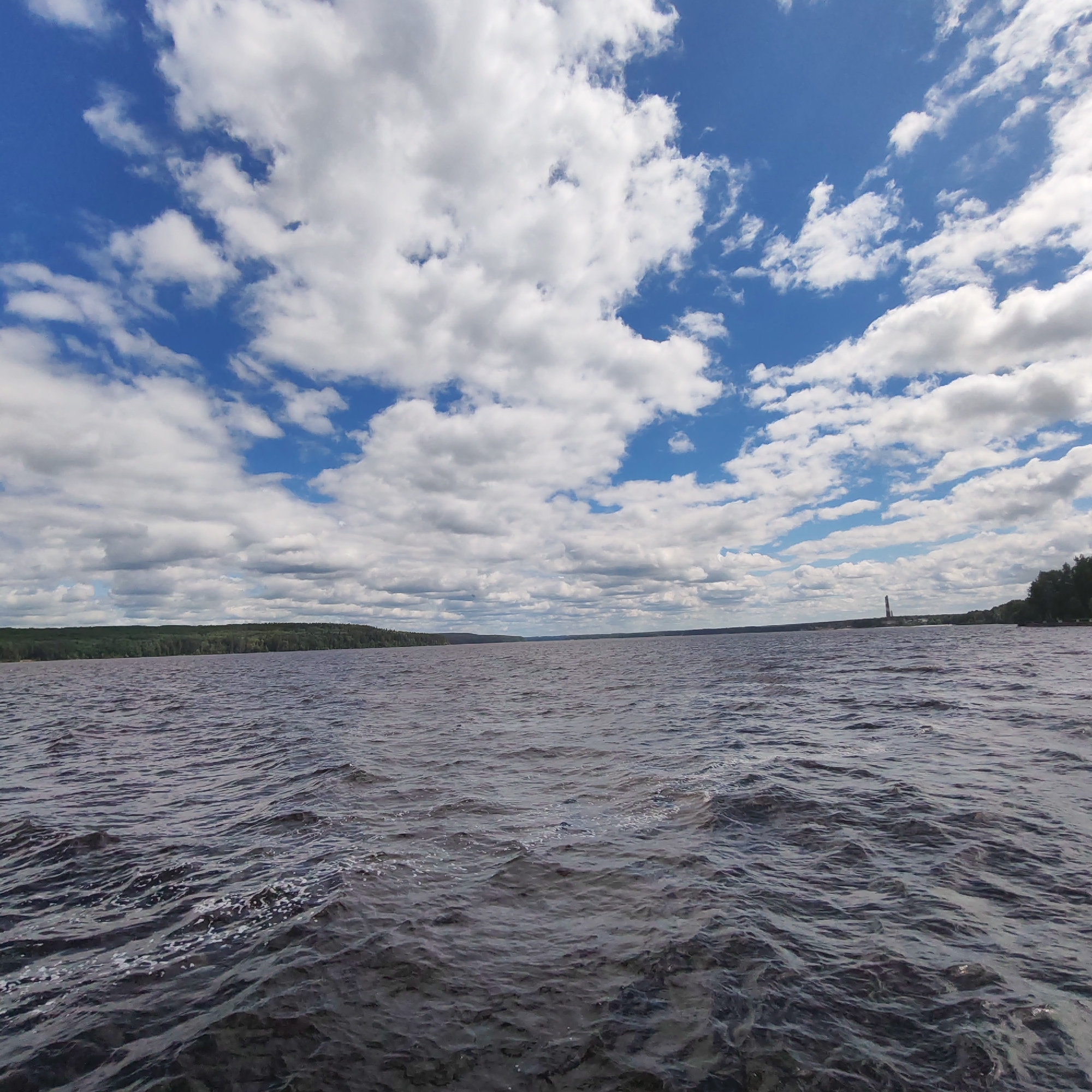 An expansive view of the Kama River under a bright blue sky filled with white, puffy clouds. The water's surface is rippled with a dark, textured pattern, suggesting a gentle breeze. Along the horizon, a dense, dark green treeline stretches across the landscape, indicating a forested shore. In the distance, a tall, slender lighthouse with red and white bands stands prominently against the sky, situated on the far bank of the river. The overall scene depicts a serene, natural environment. The image is located in the city of Chaikovsky, Russia, known for its large reservoir on the Kama River.