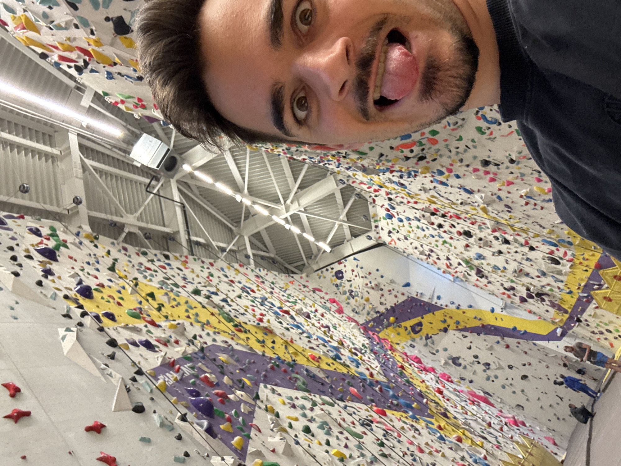 The image captures a selfie of a man with brown hair and a short beard. He has his tongue sticking out. The background shows an indoor climbing wall with various colored climbing holds. A building ceiling can be seen as well. The location is likely a climbing gym in Sofia, Bulgaria. There are other people in the background next to the climbing wall.