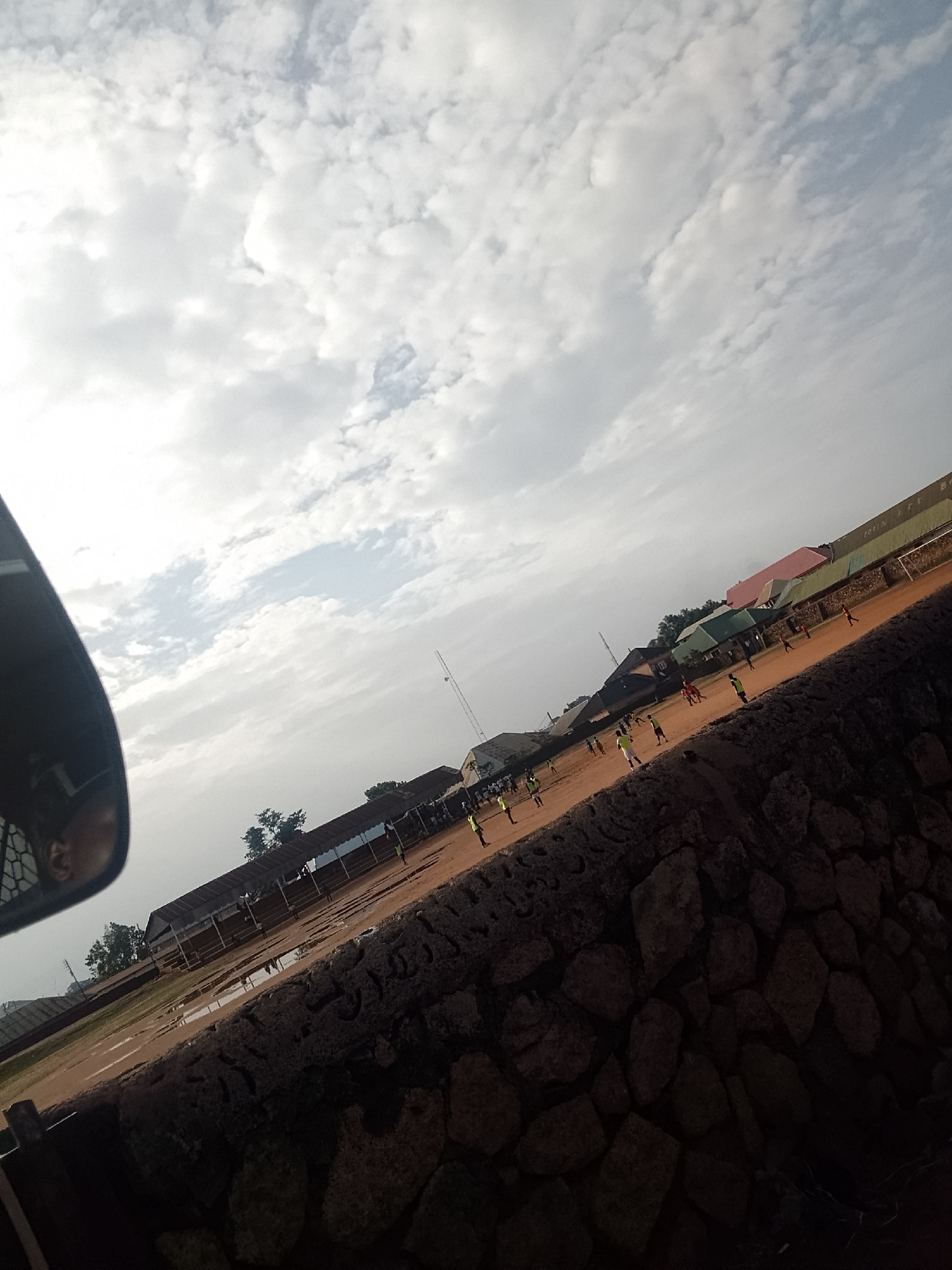The image shows a soccer field in Bukuru, Nigeria, with a match in progress. The field is surrounded by low rock walls and buildings, some with covered spectator areas. Players in green and red jerseys are visible on the field. The sky is overcast with many clouds. A portion of what appears to be a vehicle's side mirror is in the foreground on the left. The lighting is somewhat dim and muted.