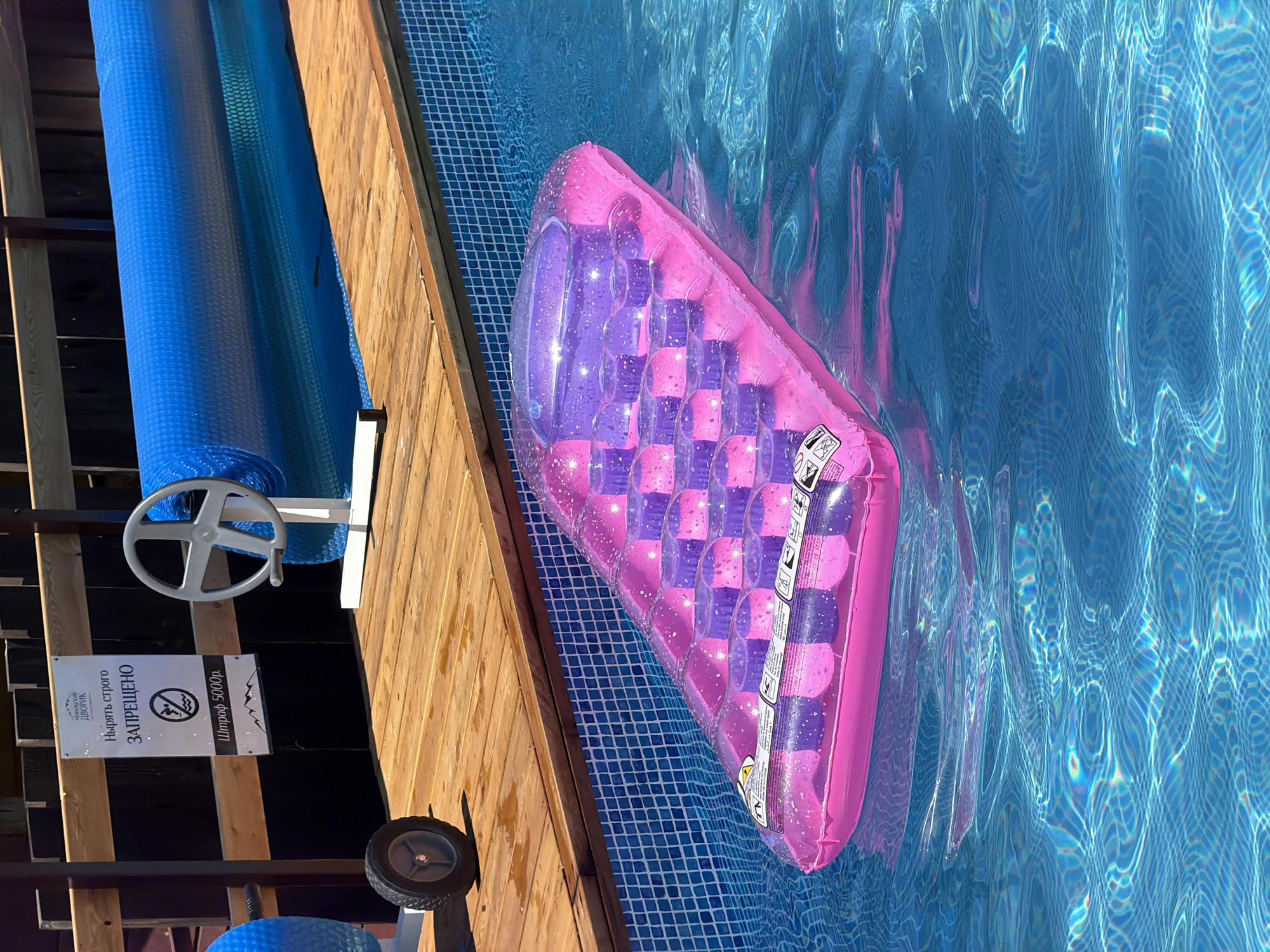 An outdoor swimming pool with blue tiled walls and clear blue water is partially covered by a rolled-up blue solar pool cover. A wooden deck surrounds the pool. A pink and purple inflatable pool float rests on the water. Next to the pool, a sign in Russian states "DO NOT DIVE - STRICTLY PROHIBITED" and indicates a fine of 5000 rubles. A wheelbarrow with a black tire is on the deck, and a large silver wheel is mounted on a white stand, near the pool cover. The background features a wooden fence and a dark structure. The location is identified as Elekmonar, Russia.