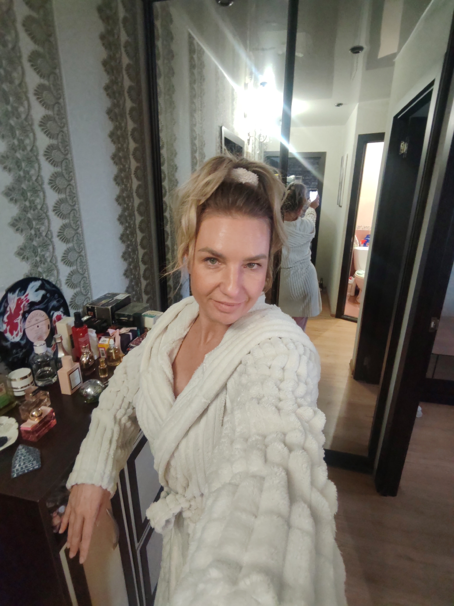 A woman in a white, textured bathrobe is taking a selfie in what appears to be a bedroom or dressing area in Petropavlovsk-Kamchatsky, Russia. Her blonde hair is tied up with a white scrunchie, and she has a relaxed, pleasant expression. To her left, a wooden dresser or vanity is visible, cluttered with numerous perfume bottles, cosmetic containers, and a red bottle, as well as a decorative vinyl record featuring a red and white design.
Behind her, a large mirror reflects a hallway, where her own back is visible as she holds a phone up to take the picture. Further down the mirrored hallway, an open doorway reveals a bathroom with a white toilet and a blue container. The hallway ceiling is reflective, showing recessed lighting. The room's wallpaper on the left features a light-colored intricate pattern, and the flooring is light-toned wood. No legible text is visible in the image.