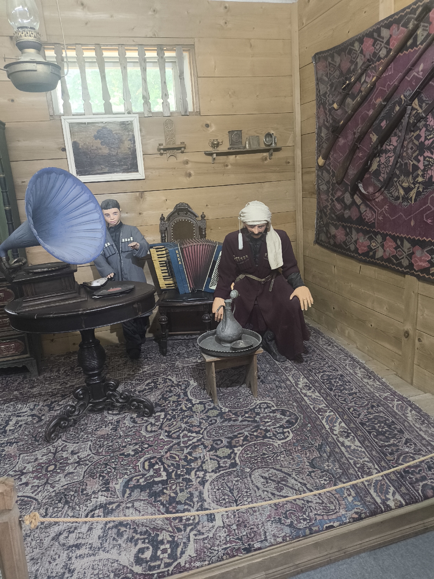 This image depicts a diorama inside a wooden room, likely a museum exhibit, located in the city of Batumi, Georgia. The room has wooden walls and floor, with a rug covering the floor area.
In the foreground, a dark wooden table with an ornate base stands prominently. On the table, there are records and a silver-colored object. A large, blue gramophone horn extends from the left side of the frame, attached to a gramophone.
Two mannequins are present. To the left, a young man mannequin is dressed in a gray traditional outfit with military-style buttons. He is positioned behind the table. To the right, a seated older man mannequin wears a dark red robe, a white headscarf, and a white cloth draped around his neck. He has a dark mustache and a stern expression. He is seated on the floor next to a small wooden stool.
On the stool, there is a metal jug and a circular tray. Behind the older man mannequin, an accordion is visible, resting on a wooden chair with an upholstered back.
The back wall of the room features several items. A large, patterned rug with red, black, and white designs hangs on the right side, with several long firearms leaning against it. To the left, a shelf holds small decorative objects and clocks. A framed, dark-colored painting is mounted on the wall above the gramophone. A window with wooden bars is visible in the upper left portion of the room, showing some greenery outside. A kerosene lamp hangs from the ceiling in the upper left corner.
