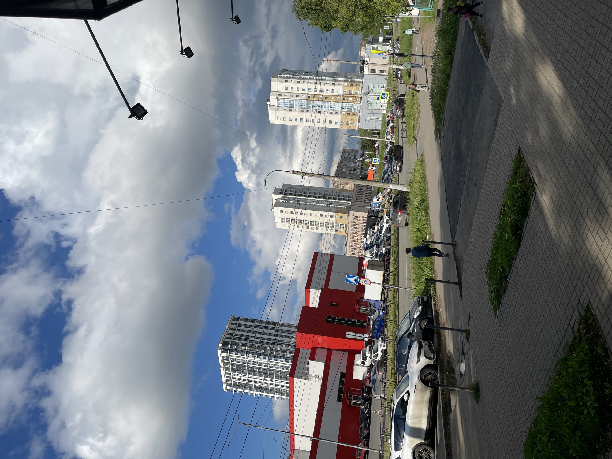 The image, taken in Izhevsk, Russia, captures an urban street scene on a bright, partly cloudy day, with the perspective rotated 90 degrees counter-clockwise. The sky features a mix of deep blue and large, dramatic white and grey cumulus clouds.
In the midground and background, several tall, modern residential buildings with light-colored facades and numerous windows dominate the cityscape. In front of one such residential tower, a prominent red and white commercial building, likely a large store or hypermarket, stands out. Faint text on its side appears to read "ЛЕНТА" (LENTA), a Russian retail chain.
A paved road runs through the scene, lined with numerous parked cars of various colors, including white, black, blue, and silver. Adjacent to the road on the right is a wide, patterned sidewalk, bordered by narrow strips of grass and small bushes. A person wearing a blue jacket is visible walking away from the viewer on this sidewalk. Other pedestrians can be seen further in the distance.
Overhead, multiple streetlights on poles and a complex network of power lines and cables crisscross the sky. A blue circular road sign with a red border and the number "30" indicating a speed limit is visible near the commercial building. The bright light and clear shadows suggest it is late morning or early afternoon.