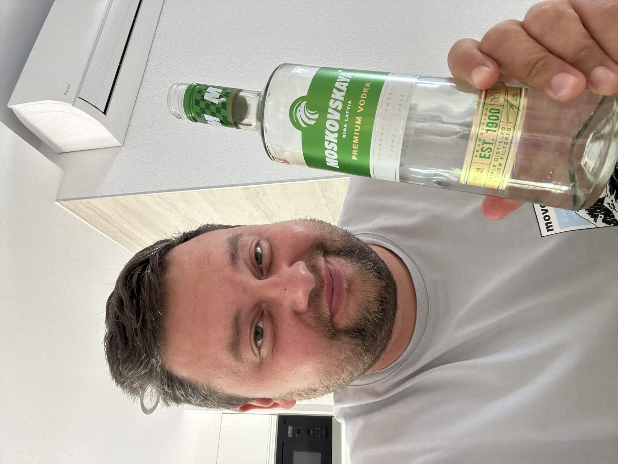 The image shows a man holding an empty bottle of Moskovskaya Premium Vodka. The man has short brown hair with a beard. He is wearing a light gray t-shirt. The vodka bottle has a green label with white lettering. The label also includes the text "Riga Latvia" and "Production Est. 1900." The background includes a white wall, a Daikin air conditioning unit, and a partially visible black oven. The location is not specified in the provided image or text.
