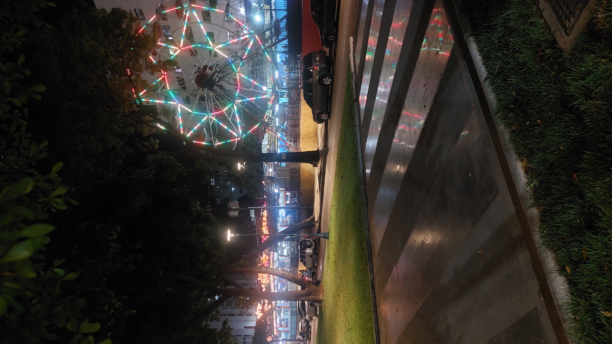 The image shows a night scene in Caracas, Venezuela, featuring a Ferris wheel illuminated with red, green, and blue lights. It is located near a street with parked cars and streetlights. The foreground includes a sidewalk with a herringbone pattern and grass. Trees partially obscure the Ferris wheel. In the background, there are buildings, also illuminated with lights.