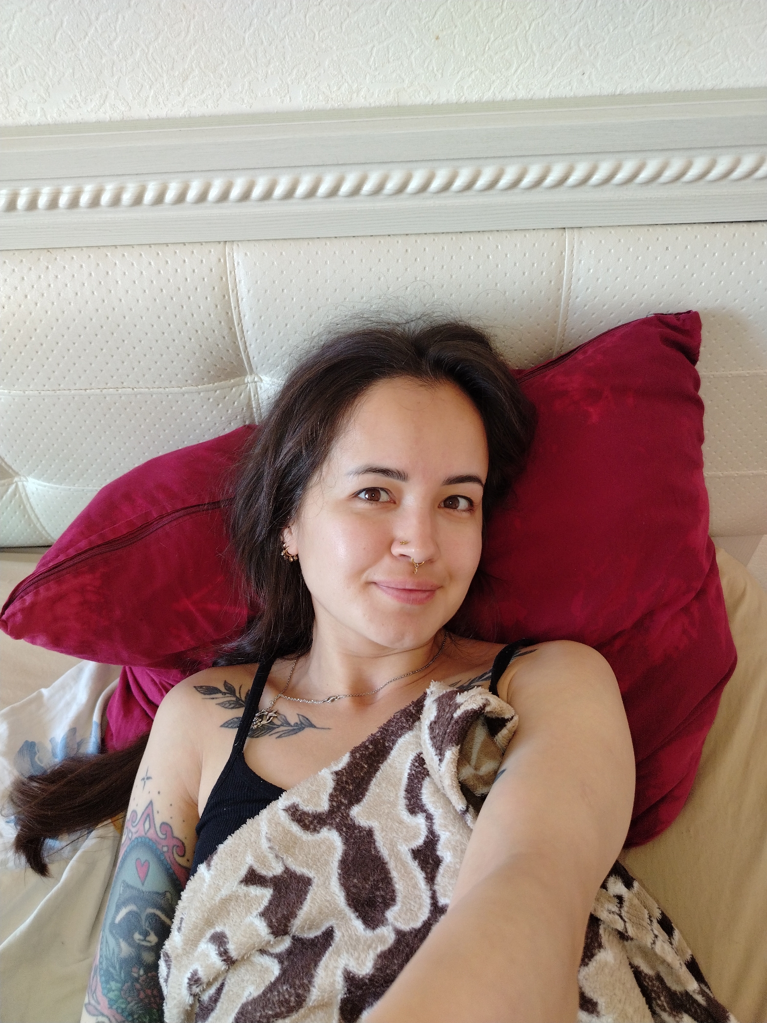 A young woman is lying in bed, resting her head on a maroon pillow. She is looking at the camera with a slight smile. She has long, dark hair and is wearing a black tank top. Her left arm is covered by a blanket with a brown and white animal print pattern. She has several tattoos on her arms and shoulder. The bed has a white headboard with a textured pattern. The room appears to be a bedroom. The image does not provide any information about the location in the city of Noviy, Russia.