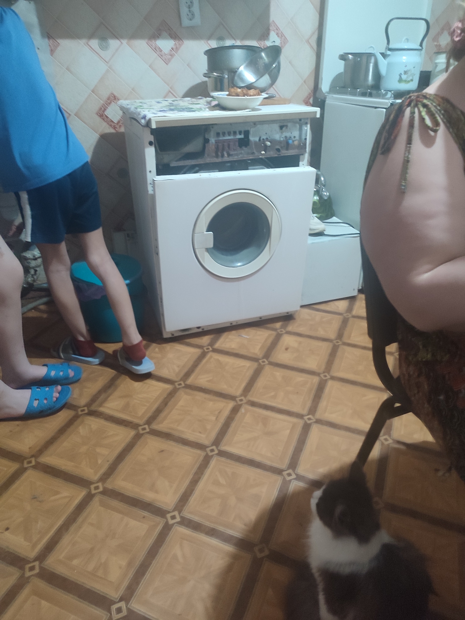 The image depicts a domestic scene, likely a kitchen, with a partially disassembled washing machine in the foreground. To the left, a person's legs are visible, wearing blue slide sandals and dark shorts. To the right, another person's torso is visible, wearing a patterned dress or garment. A cat is also present in the lower right corner, looking upwards. The environment features tiled flooring and a tiled backsplash on the wall. Appliances such as a stove and a kettle are visible in the background. The image appears to be taken indoors, possibly in an apartment or house. There is no information within the image that indicates a specific location in Chernivtsi, Ukraine.