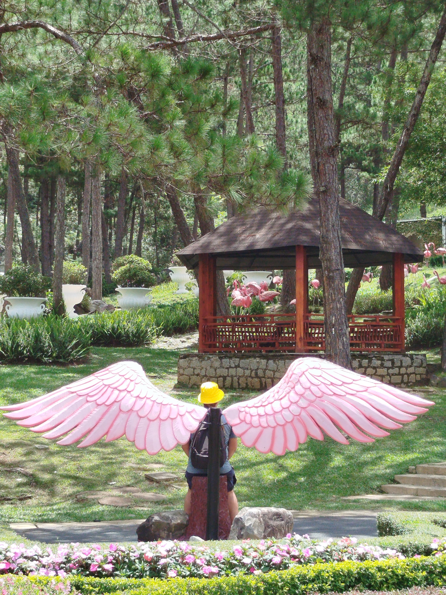 The image depicts a person wearing a backpack and a yellow hat, standing in front of a large pink wing installation in a park. In the background, a wooden gazebo with a dark roof is visible, surrounded by trees and lush greenery. Pink flamingo statues are scattered throughout the park, some near the gazebo and others in the distance. The park is characterized by tall pine trees, manicured bushes, and flowering plants in the foreground. The ground is covered in grass, with stone pathways and steps visible in some areas. The overall impression is that of an outdoor recreational area or a themed attraction, likely a tourist spot in the city of Lâm Viên - Đà Lạt, Vietnam.