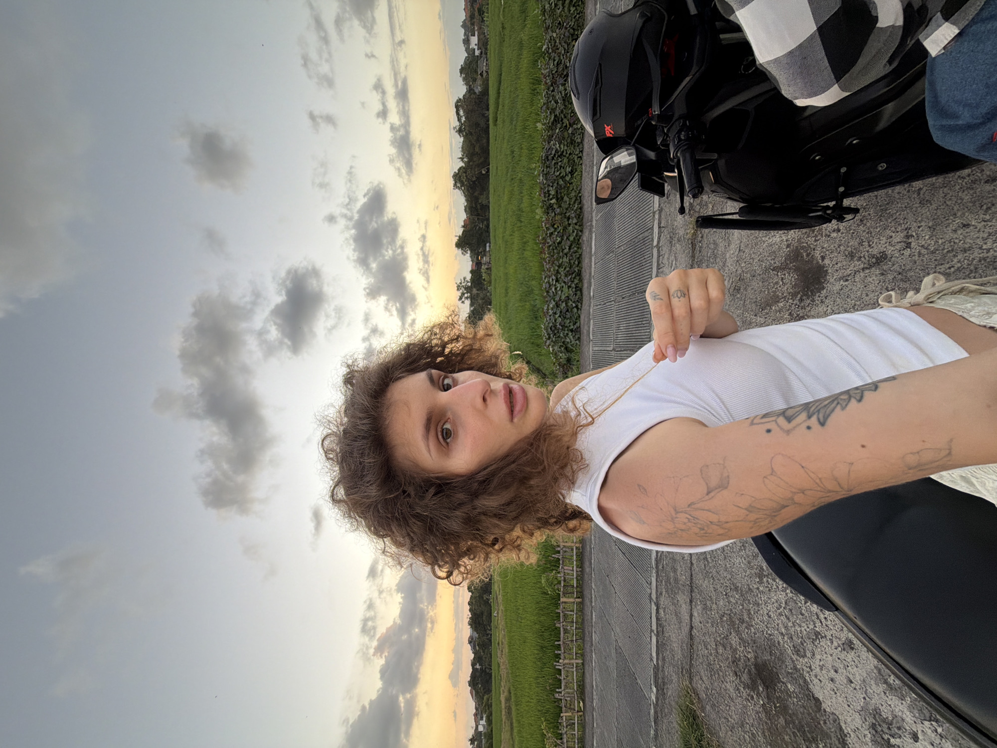 A selfie shows a young adult with curly brown hair and tattoos on their arm, wearing a white tank top and light-colored pants. They are standing next to a black motorcycle with a helmet on the handlebars. In the background, a lush green rice field is visible under a cloudy sky with the soft glow of sunset. The scene appears to be in a rural area on the outskirts of North Kuta, Indonesia.