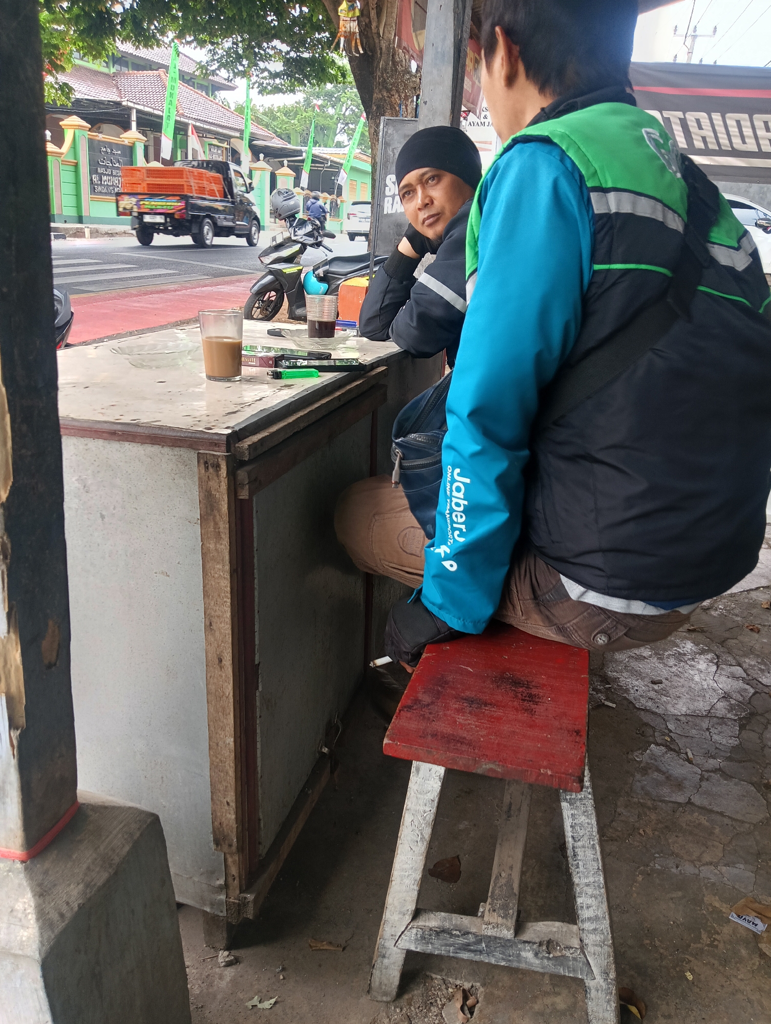 Two men are sitting at a counter in Manggari, Indonesia. The man on the left is wearing a black beanie and a dark jacket, and is looking to the left. The man on the right is wearing a blue and green jacket and is looking away from the camera. On the counter are two glasses of drink, a pack of cigarettes, a lighter, and some pens. A motorcycle is parked behind the men. In the background, a black pickup truck is driving on the road, and there are buildings with green and white banners. The ground is concrete and dirt.