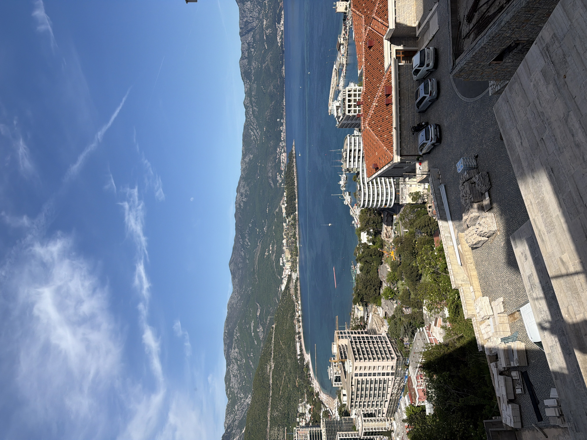 The image presents a high-angle, panoramic view of a vibrant coastal city, bay, and dramatic mountainous landscape under a bright blue sky. The scene is dominated by the deep blue expanse of the sea, which stretches across the midground, featuring several boats and yachts, some docked in a marina and others sailing in the open water. A sandy beach curves along the coastline, bordered by lush green trees and a mix of buildings.
In the foreground, on the right side of the frame, are several urban structures. These include buildings with traditional red-tiled roofs alongside more modern, multi-story white buildings, some appearing to be under construction with cranes visible. A paved area, likely a street or courtyard, is visible, featuring several parked white or silver cars and stacks of building materials (slabs of stone or tiles), indicating ongoing construction or renovation activity. Strong sunlight casts distinct shadows across the buildings and paved areas, suggesting either mid-morning or late afternoon.
The background, on the left, is dominated by a majestic, verdant mountain range covered in green foliage, rising steeply from the coastline and extending into the distance. The sky above is clear and blue with scattered, wispy white clouds, indicative of good weather. No specific human activity or event is the central focus, but the presence of boats, cars, and construction suggests a bustling coastal environment. The characteristic geography and architecture align with the provided location context of Budva, Montenegro. There is no visible text in the image.