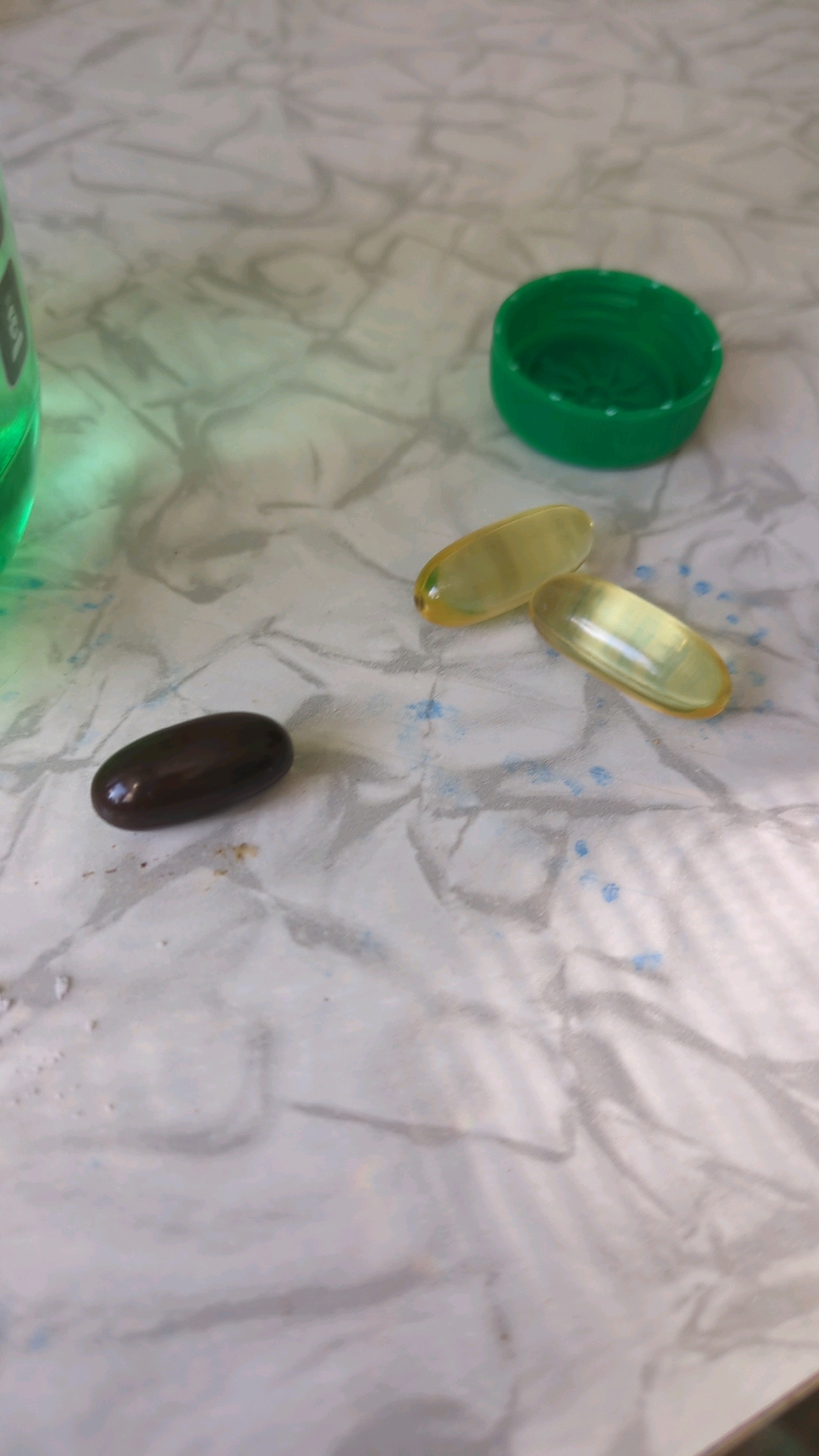 The image shows a dark brown, oval-shaped pill in the foreground, slightly to the left. To the right of the dark pill are two translucent, yellowish oval-shaped pills. A green plastic bottle cap is visible in the upper right quadrant of the image. A partially visible green bottle, likely containing a liquid, is in the upper left corner of the image. The objects are resting on a white, textured surface that appears to be crumpled paper or fabric, with some faint blue speckles scattered across it. The scene is indoors, with soft lighting. There is no information in the image to determine its location in Chișinău, Moldova.